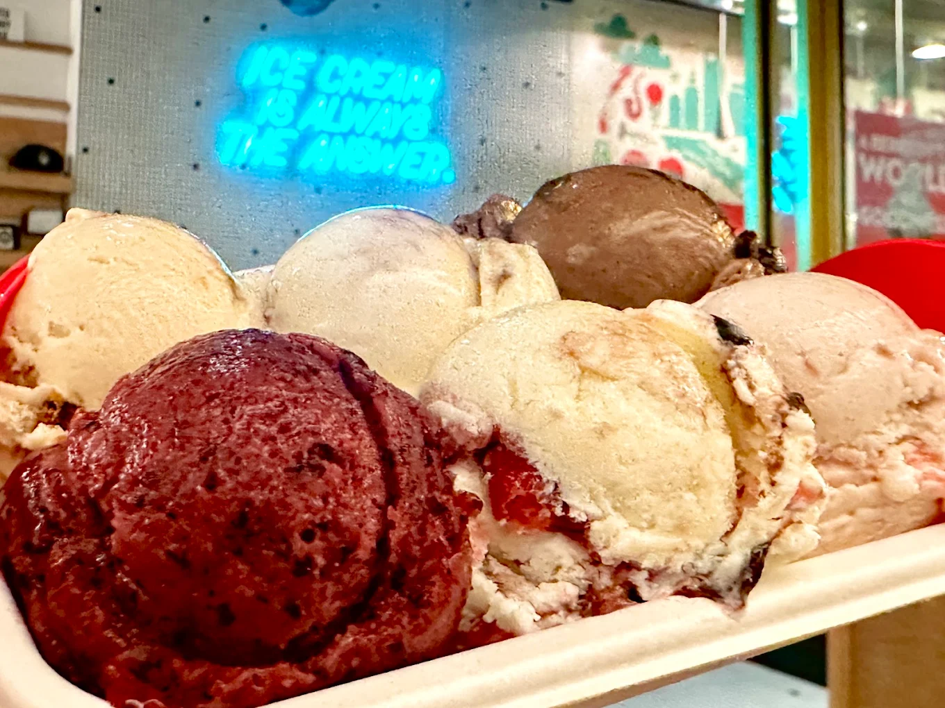 A tray holding six scoops of assorted ice cream flavors is in focus, with a neon sign in the background reading "Ice cream is always the answer." The setting appears to be inside an ice cream shop.