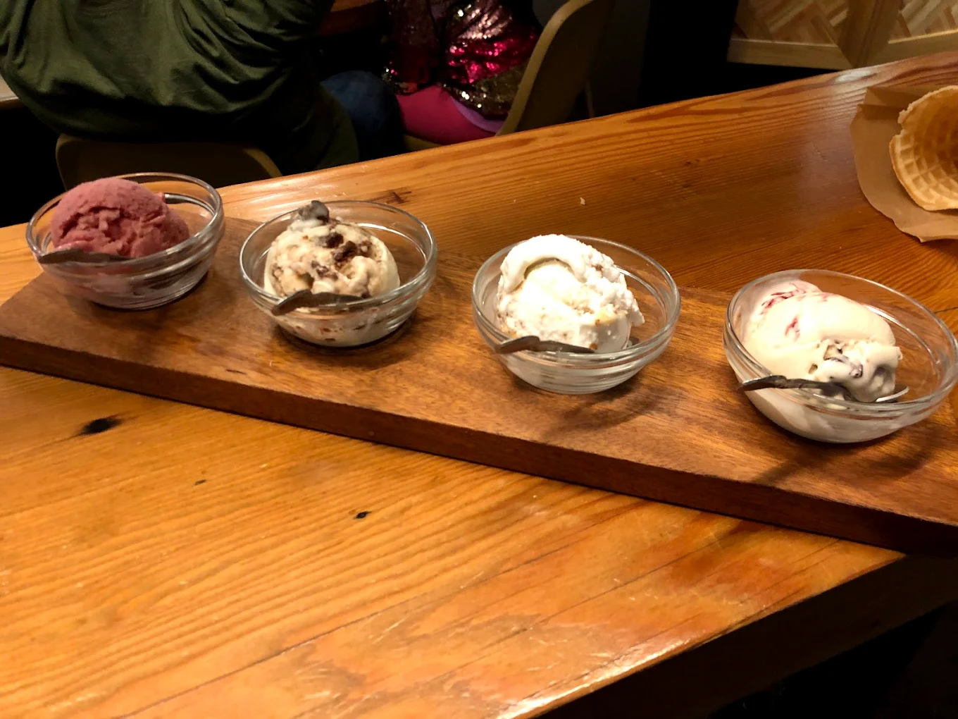 Four small glass bowls of different ice cream flavors are lined up on a wooden board, each with a metal spoon. The setting is a wooden table, and there are waffle pieces and people in the background.