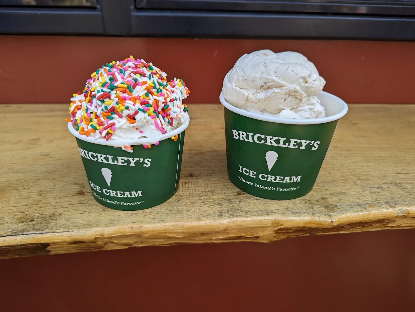 Two green cups of Brickley’s Ice Cream on a wooden ledge; the left cup has vanilla ice cream with colorful sprinkles, and the right cup has a plain scoop, likely a different flavor.