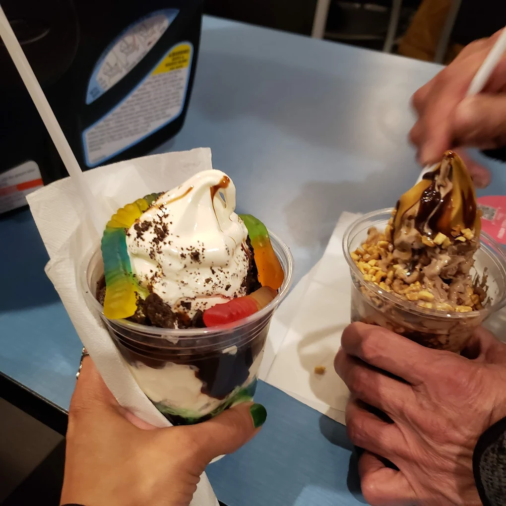 Two people holding ice cream sundaes in clear cups; one sundae has gummy worms and chocolate sauce, the other has chocolate sauce and chopped nuts. Both have spoons and are on a blue tabletop.