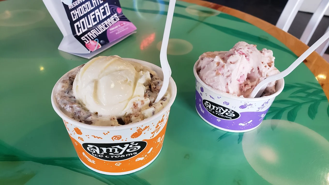 Two cups of Amy’s Ice Creams on a green table—one in an orange cup with vanilla and cookie mix-ins, and one in a purple cup with strawberry ice cream. Both have white plastic spoons. A bag is in the background.