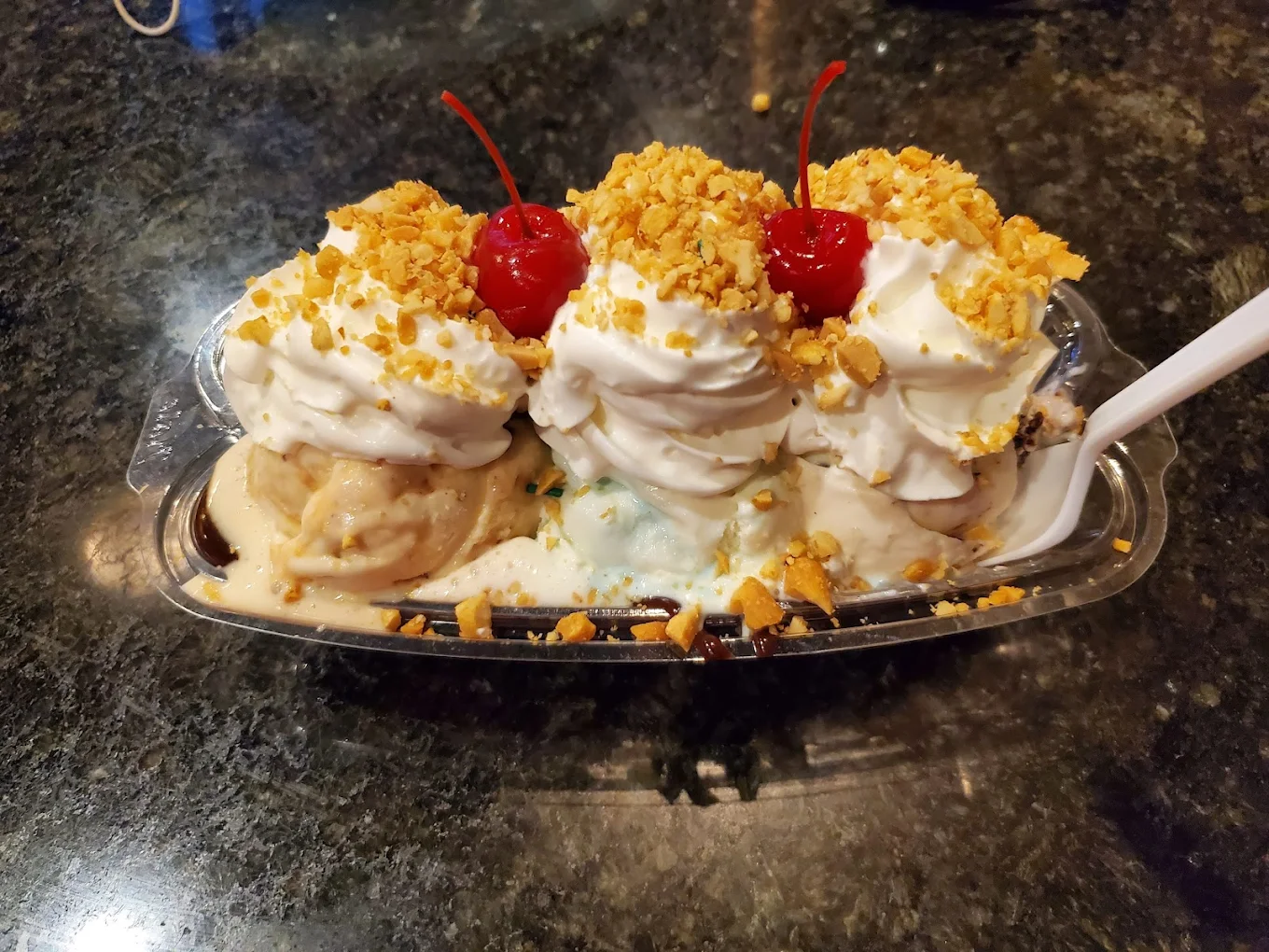 A banana split sundae with three scoops of ice cream, topped with whipped cream, chopped nuts, and a cherry on each scoop, served in a clear dish on a dark countertop.