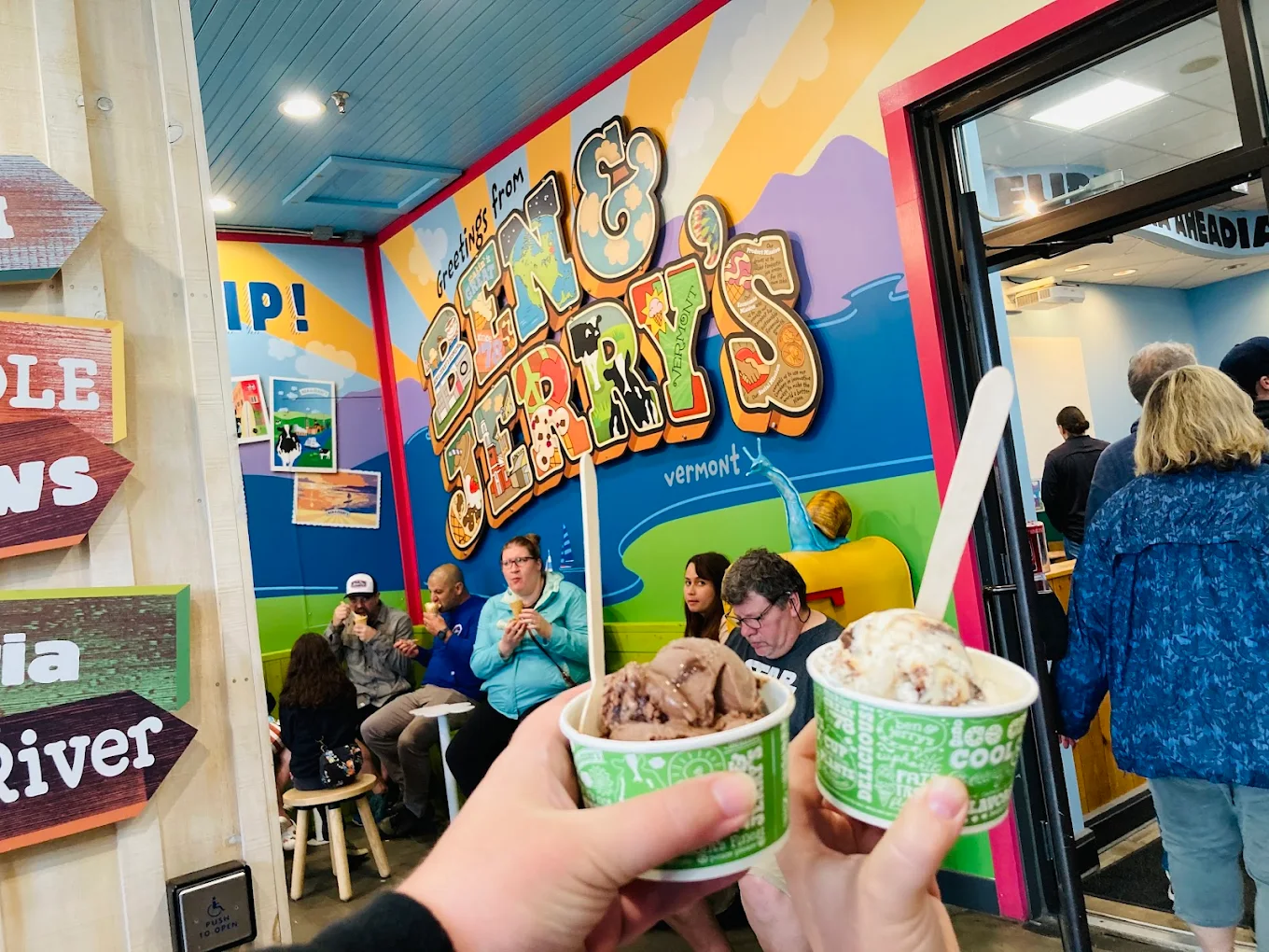 Two people hold cups of ice cream inside a brightly colored Ben & Jerry's shop, with customers seated and a large mural featuring the Ben & Jerry's logo on the wall in the background.