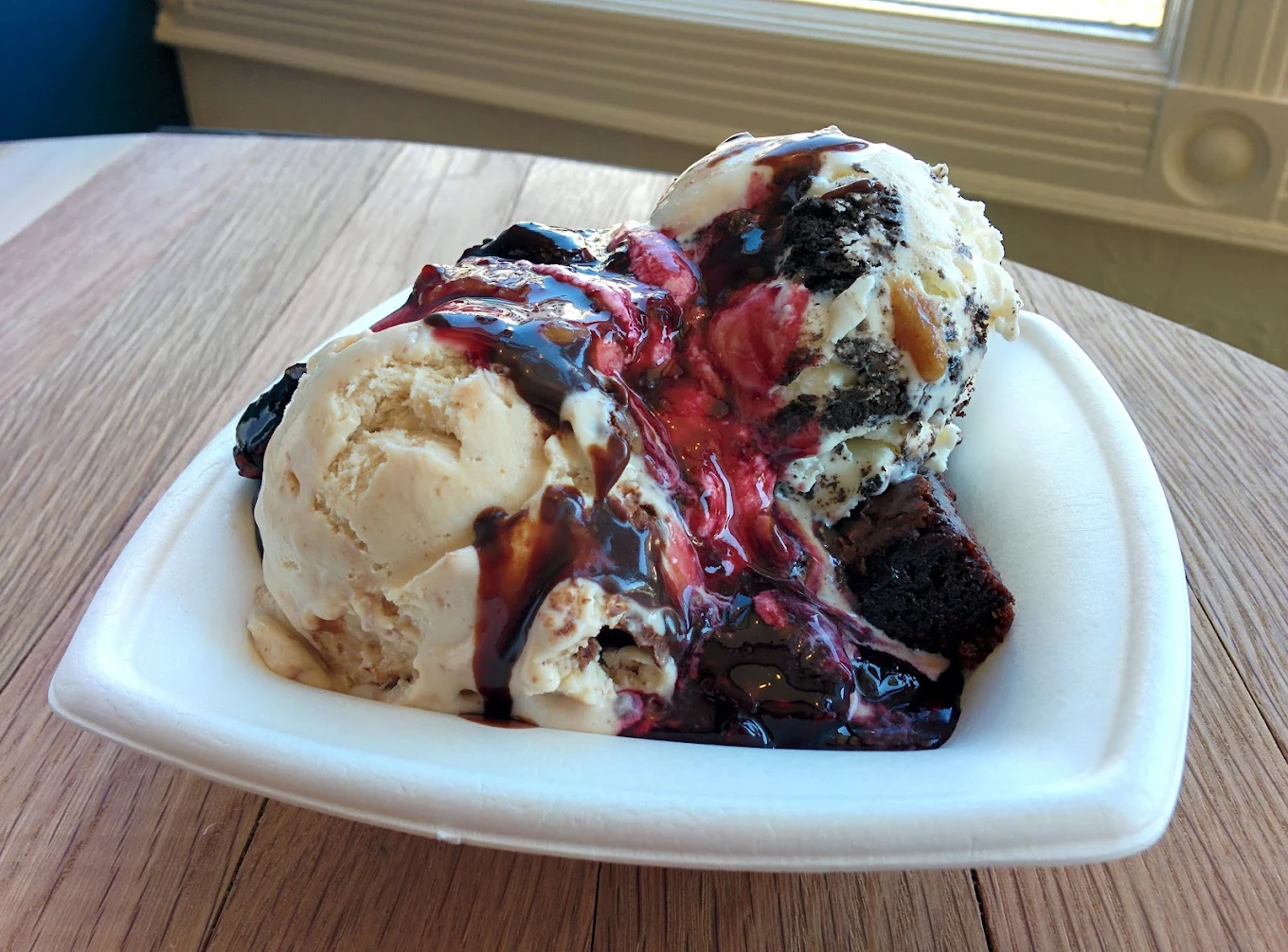 A bowl with two scoops of vanilla ice cream topped with chocolate and berry sauces, served over a brownie, placed on a wooden table near a window.