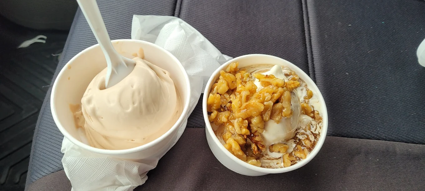 Two cups of soft serve ice cream sit on a dark car seat. The left cup has plain ice cream with a white spoon, and the right cup is topped with walnuts and syrup. Both cups are placed on napkins.