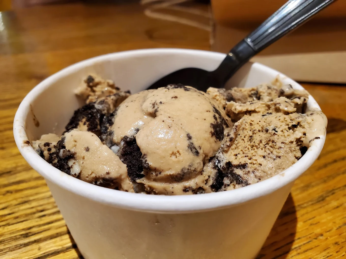 A close-up of a cup of cookies and cream ice cream with chocolate cookie chunks, served in a white paper cup with a black plastic spoon on a wooden table.