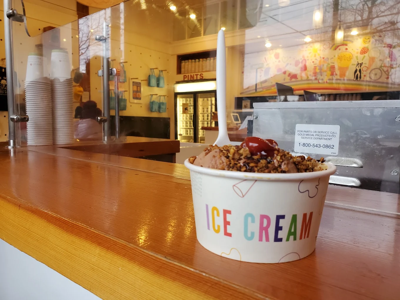 A cup labeled "ICE CREAM" filled with chocolate ice cream, nuts, and a cherry sits on a wooden counter inside an ice cream shop. The interior features shelves, cups, a pint freezer, and colorful wall art.