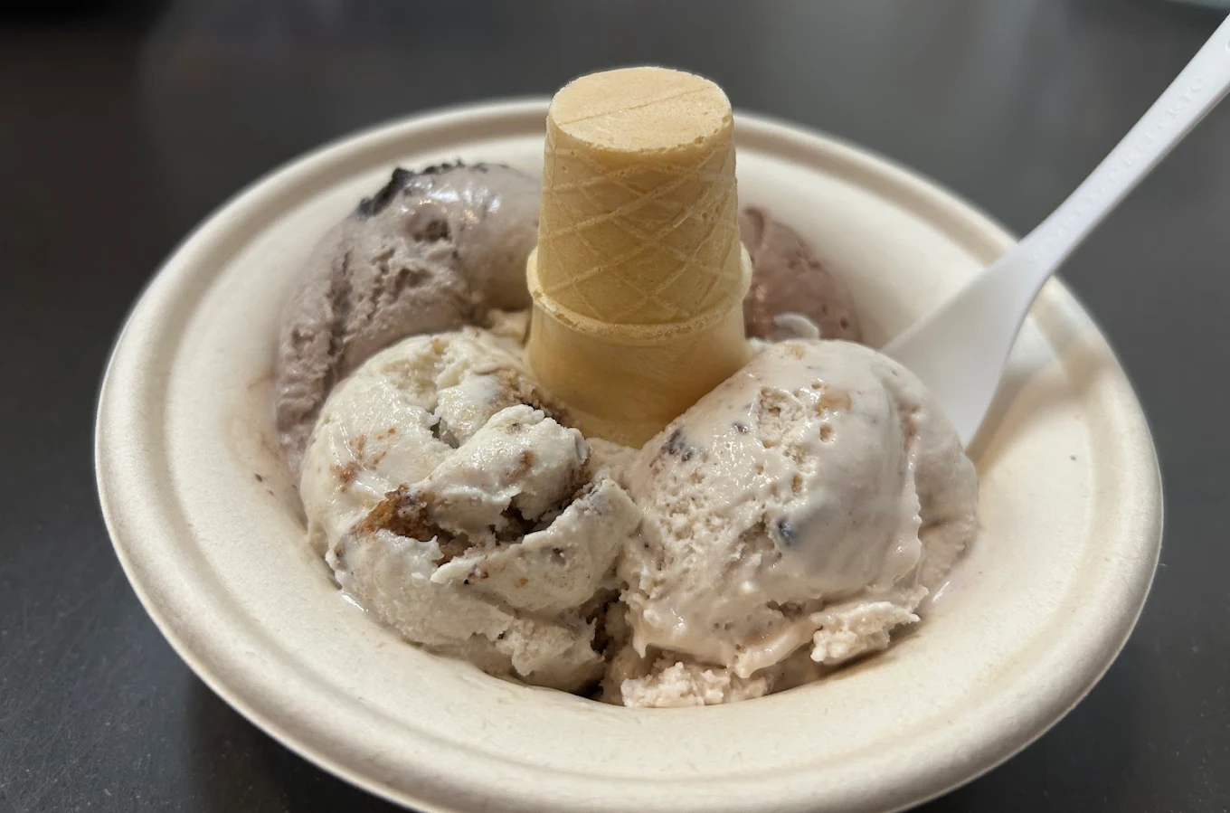 A bowl with three scoops of ice cream in different flavors, a small upside-down ice cream cone placed in the center, and a white plastic spoon on the right side.