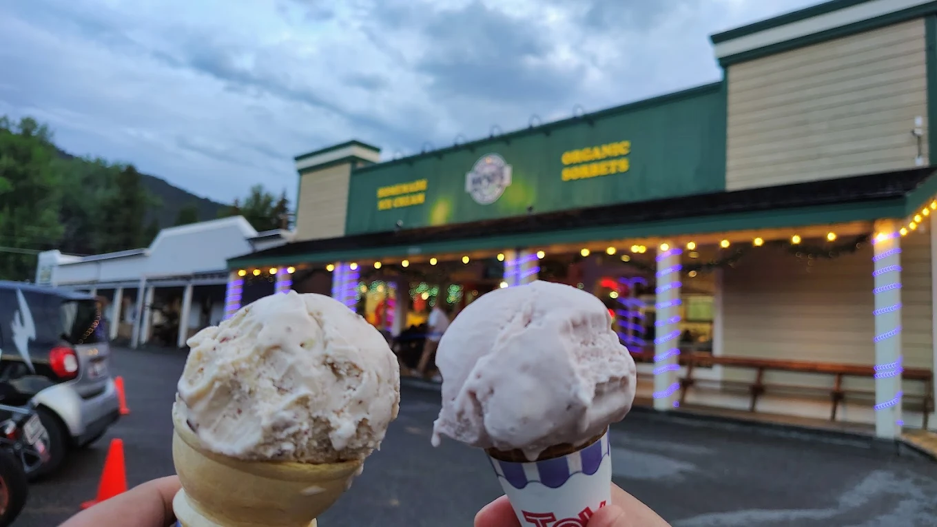 Two hands hold ice cream cones in front of a green and yellow building with a sign that reads "Organic Sweets." The sky is cloudy, and colorful lights decorate the storefront.