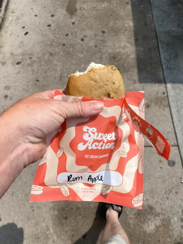 A hand holds a Sweet Action ice cream sandwich in a cookie, partially unwrapped. The wrapper has a red and white design and is labeled “Rum Apple.” The background shows a sidewalk with shadows.