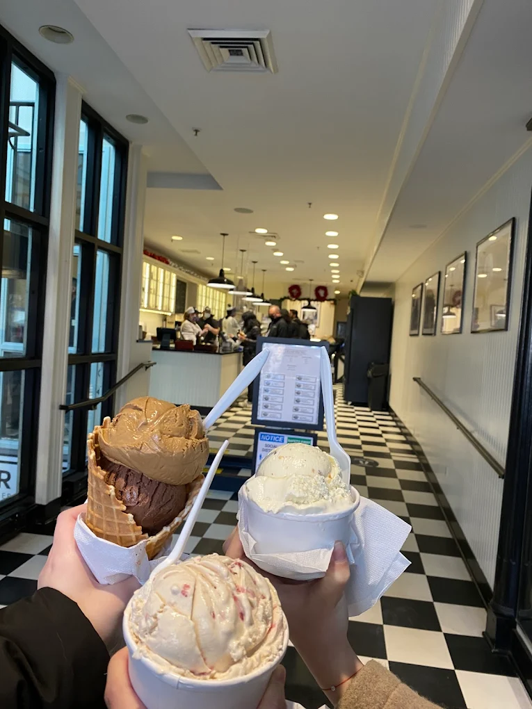 Three people hold up different ice cream cones and cups inside a bright ice cream shop with black-and-white checkered floors, large windows, and a counter in the background where people are ordering.