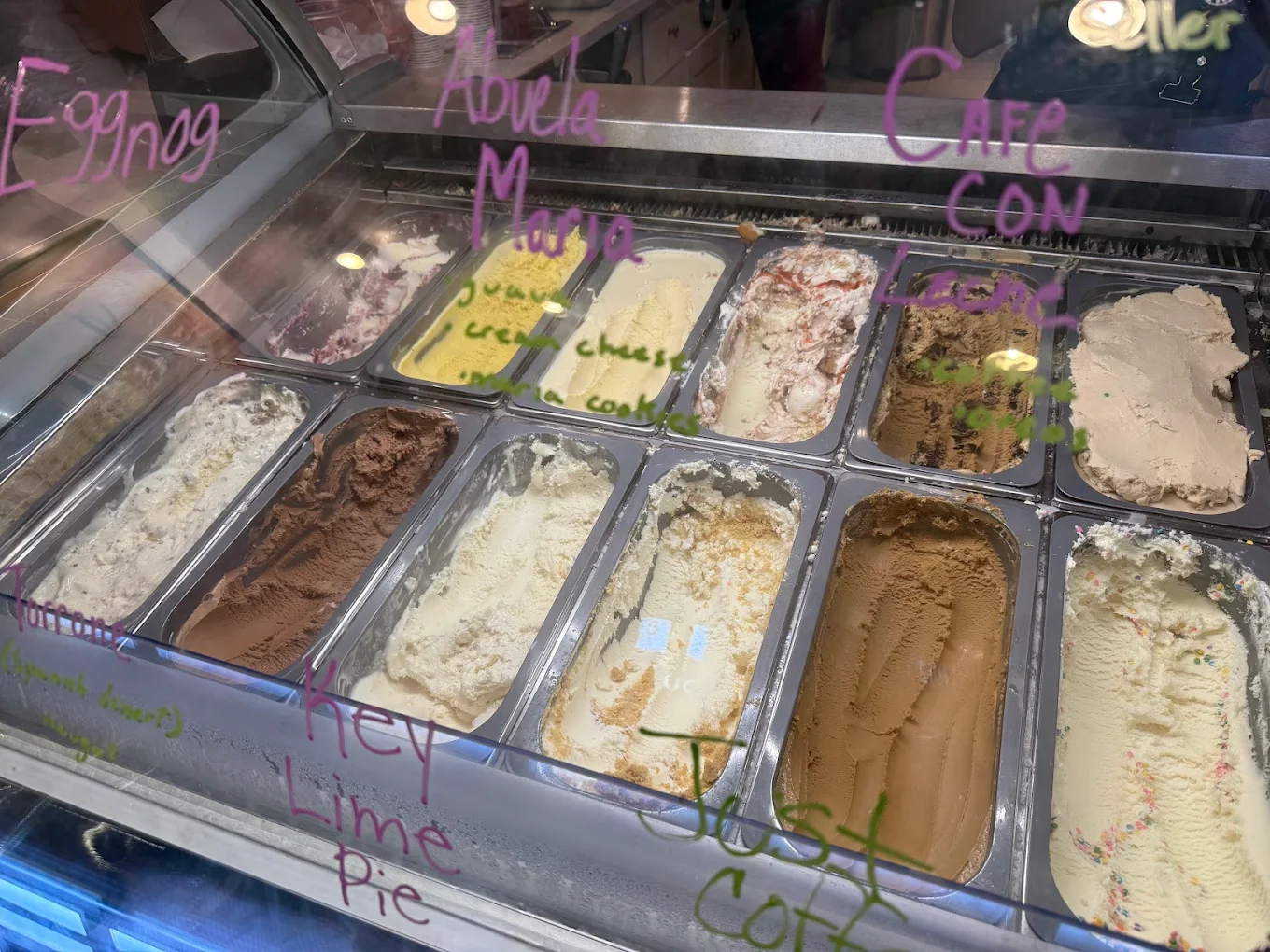 A display case of various ice cream flavors in metal trays, with flavor names like Eggnog, Adwela Mojo, Key Lime Pie, and Cafe Con Leche written in colorful marker on the glass.