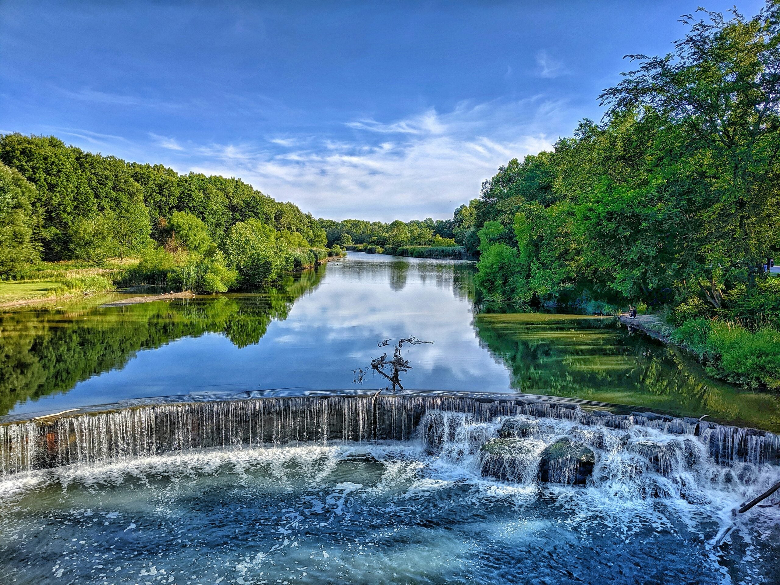 A serene river scene with a small waterfall in the foreground, lush green trees on both sides, and a clear blue sky reflected in the calm water.