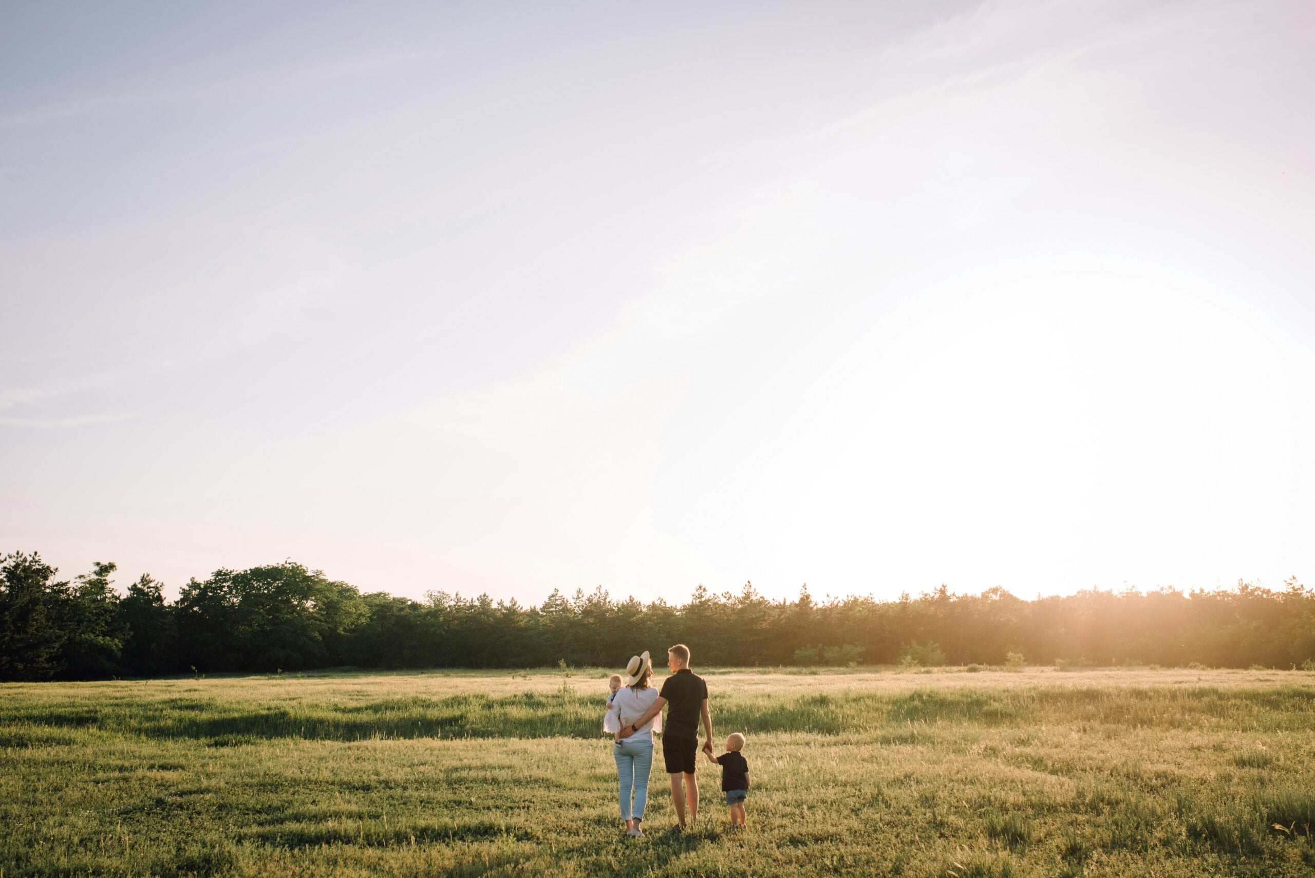 A family of three, two adults and a child, stand together in a sunlit field, holding hands and facing away, with trees in the distance and a bright sky overhead.