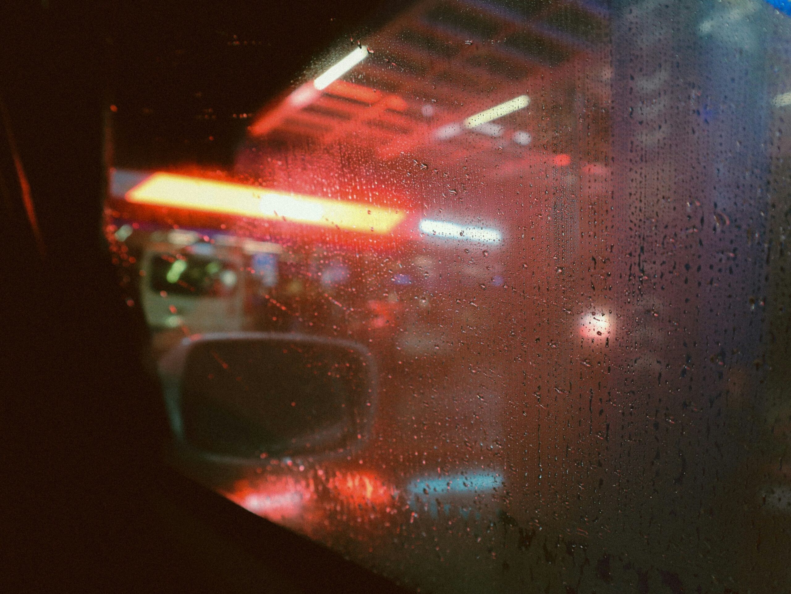 Rain-speckled car window at night with colorful, blurred neon lights from a gas station or convenience store visible outside. The scene feels moody and atmospheric.