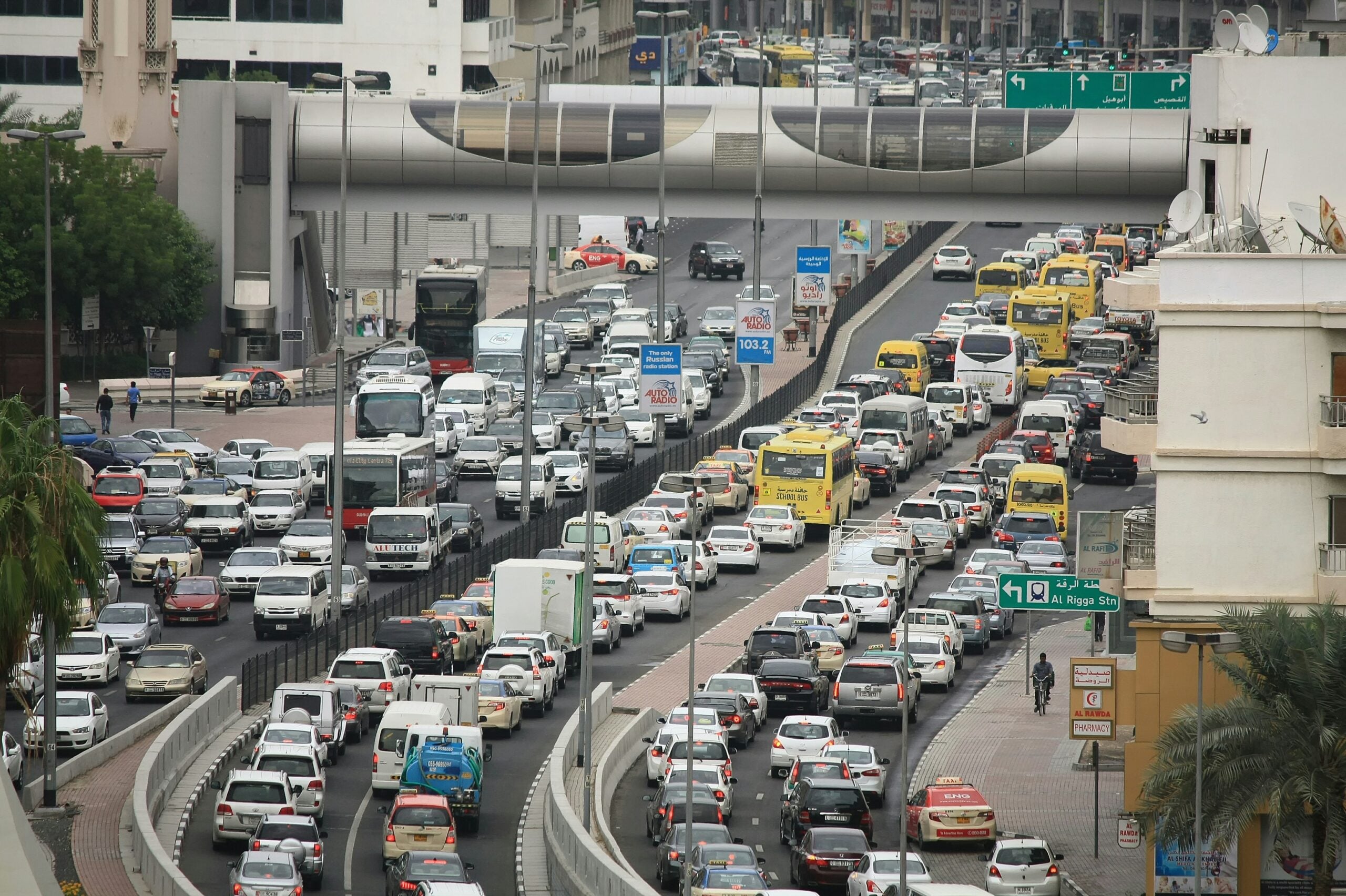 A busy urban street with heavy traffic congestion, featuring many cars, taxis, and yellow buses. Buildings and a pedestrian bridge are visible in the background. Traffic signs and palm trees line the road.