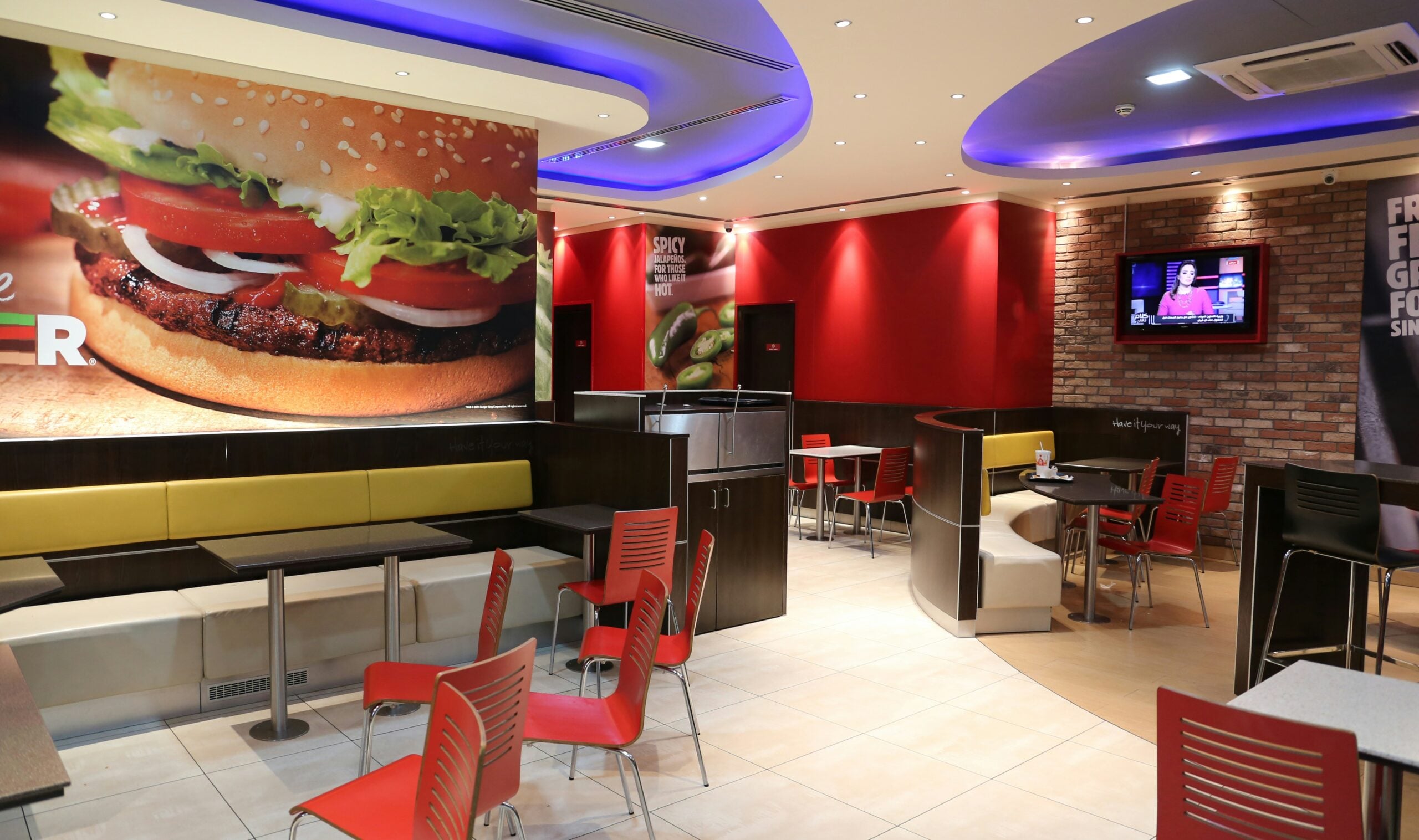 A modern fast food restaurant interior with red chairs, yellow and brown booths, empty tables, bright lighting, and a large burger poster on the wall. A TV is mounted on a brick wall in the background.
