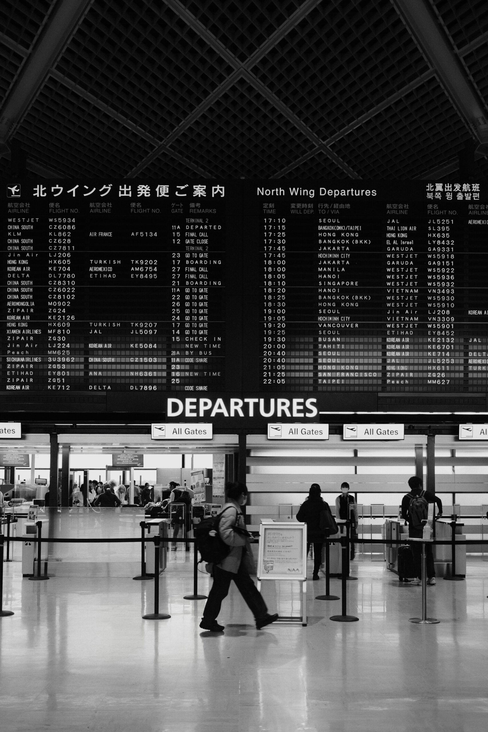 A person walks through an airport terminal under large departure boards displaying flight information in Japanese and English. Several other travelers stand near check-in counters. The scene is in black and white.