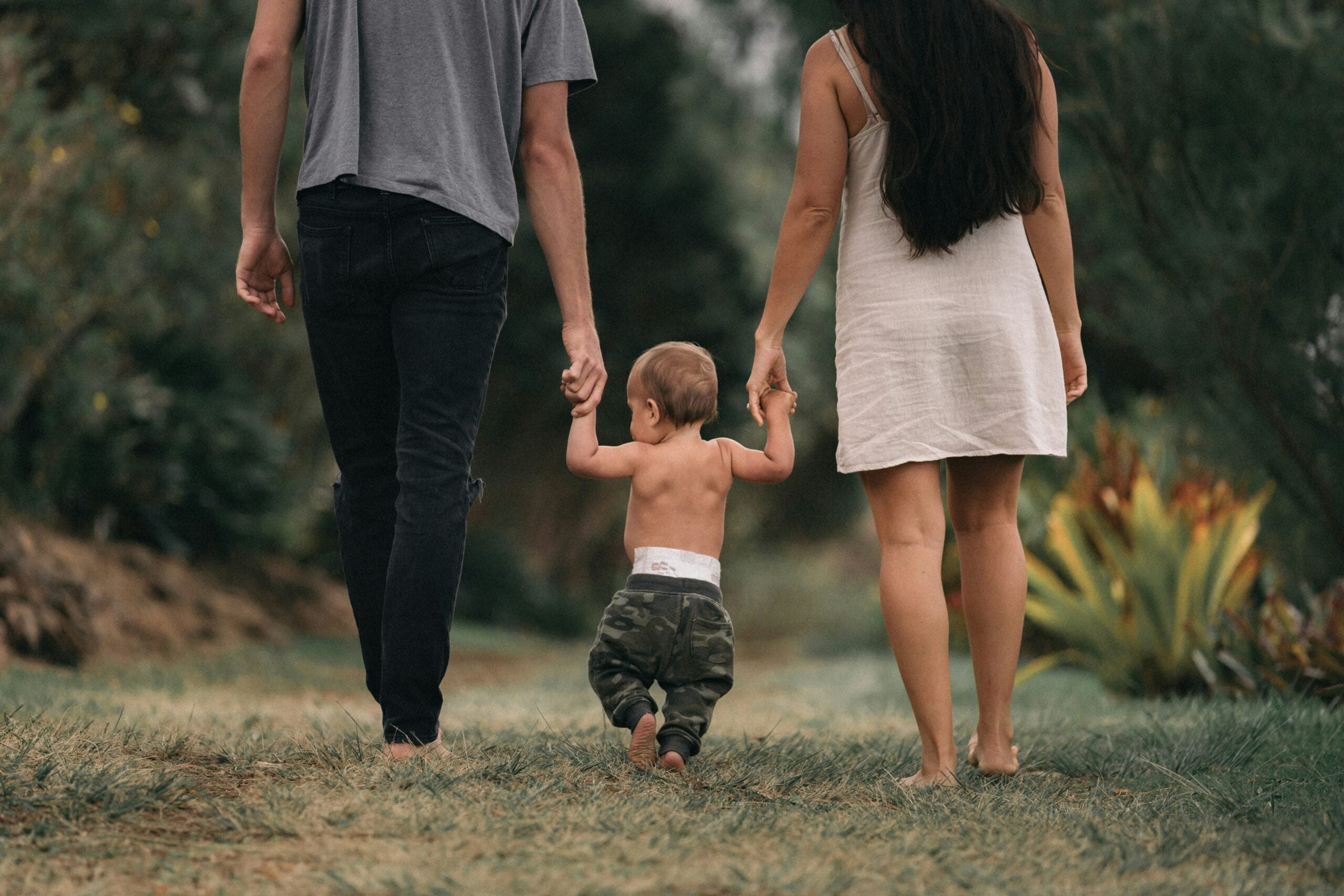 A toddler walks barefoot outdoors, holding hands with two adults on either side. The group is seen from behind, walking on grass among green trees and plants.