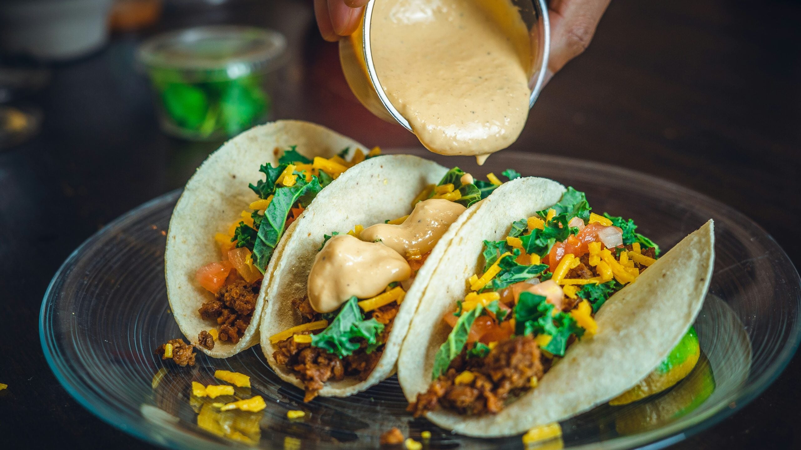Three soft tacos filled with seasoned ground meat, shredded cheese, chopped greens, and diced tomatoes are on a glass plate. A hand pours creamy sauce over the tacos. A small container of green garnish is in the background.