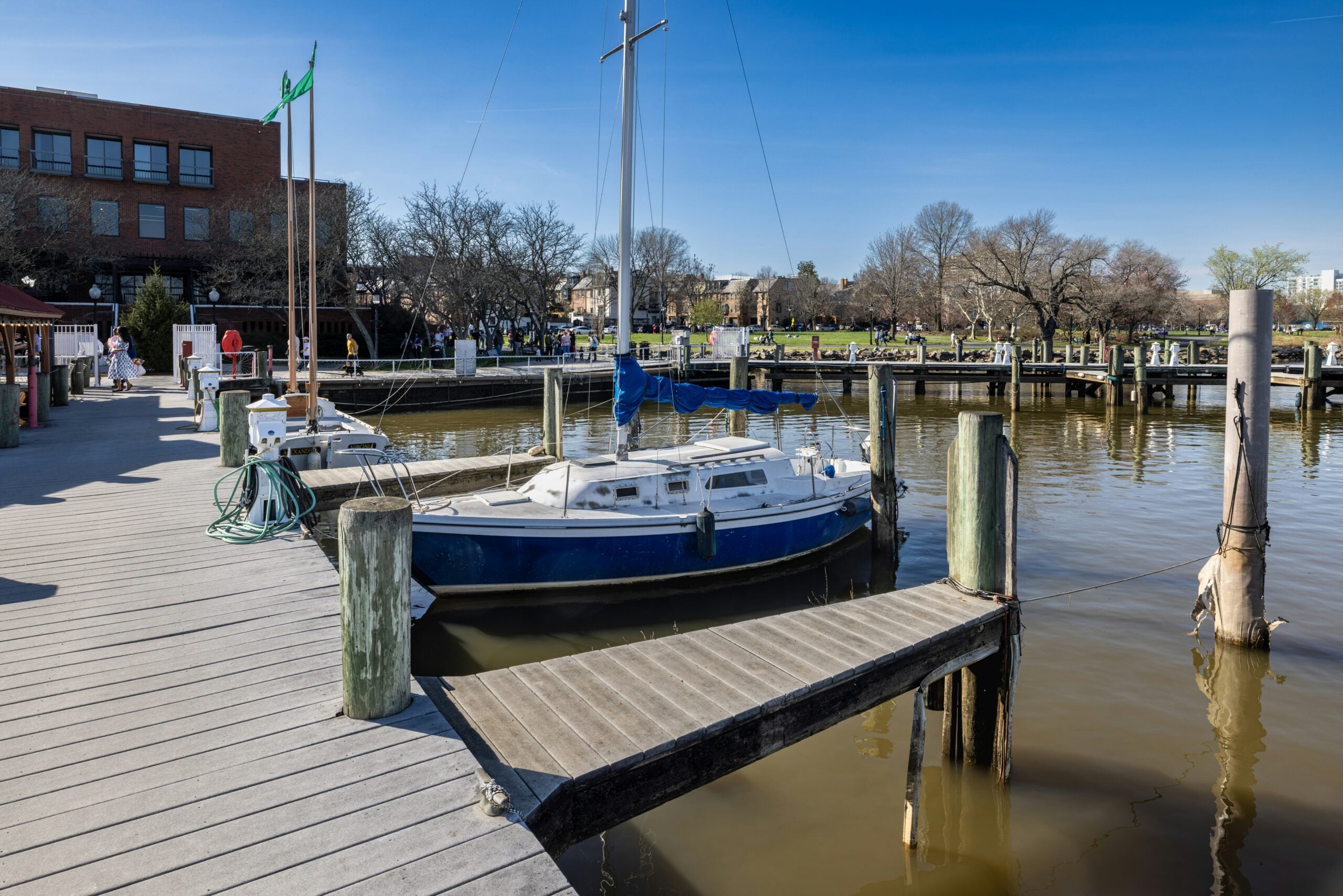 A blue and white sailboat is docked at a wooden pier on a calm river, with trees, a brick building, and people walking in the background under a clear blue sky.