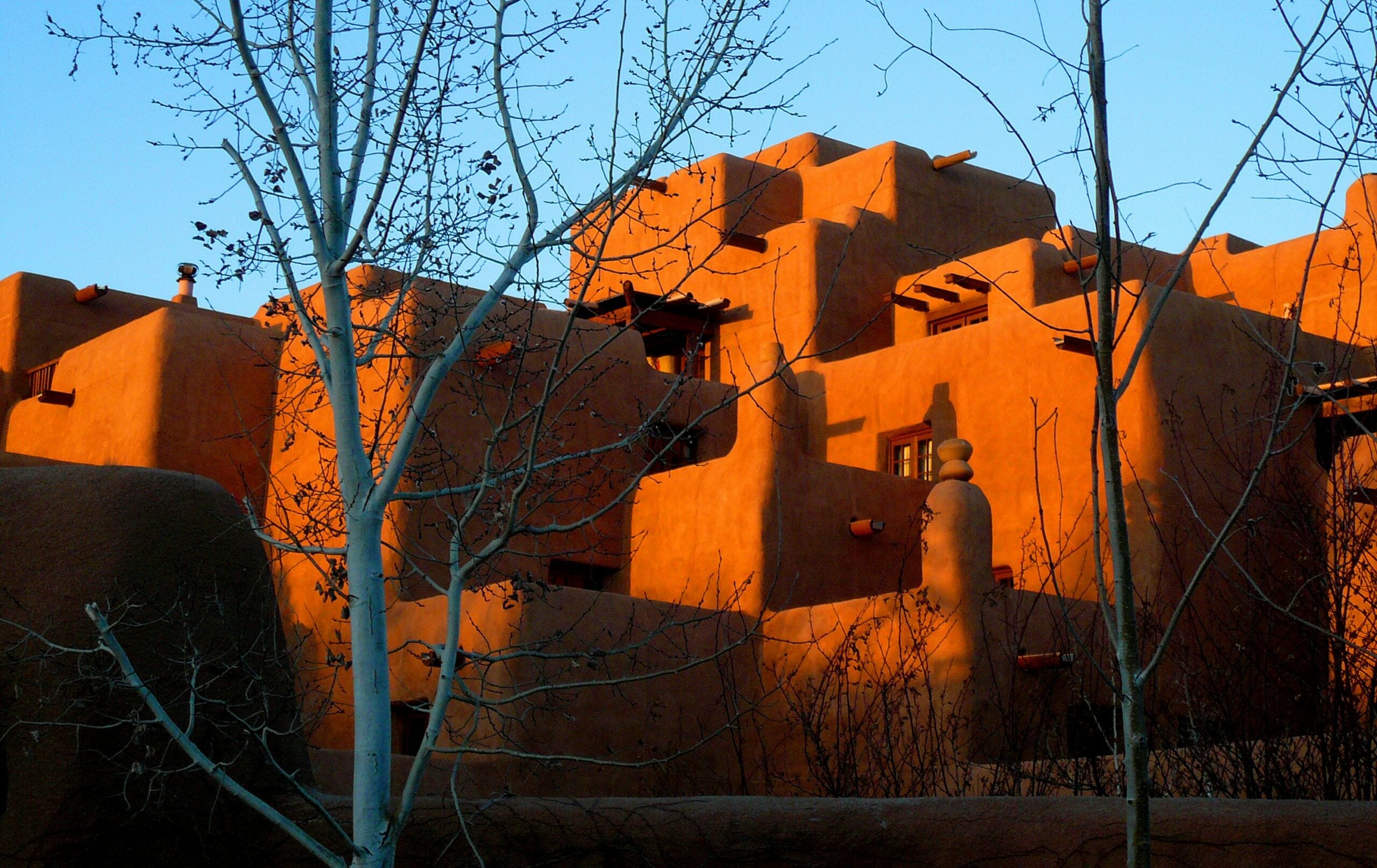 Adobe-style building with multiple levels and terraces, bathed in warm sunlight. Bare tree branches are visible in the foreground against a clear blue sky.