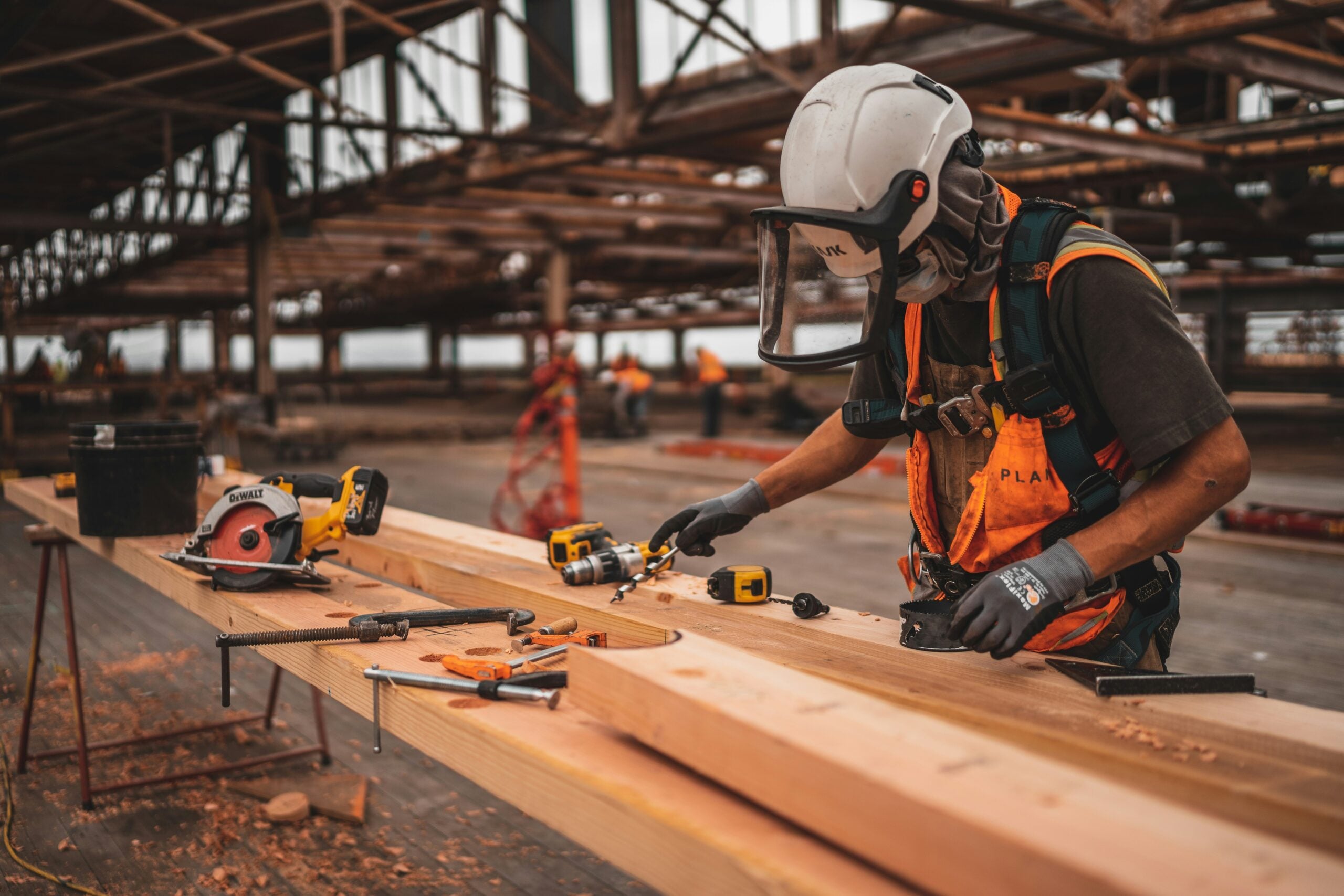 A construction worker wearing safety gear uses a power drill on wooden beams at a large indoor construction site. Various tools and equipment are spread across the workbench.