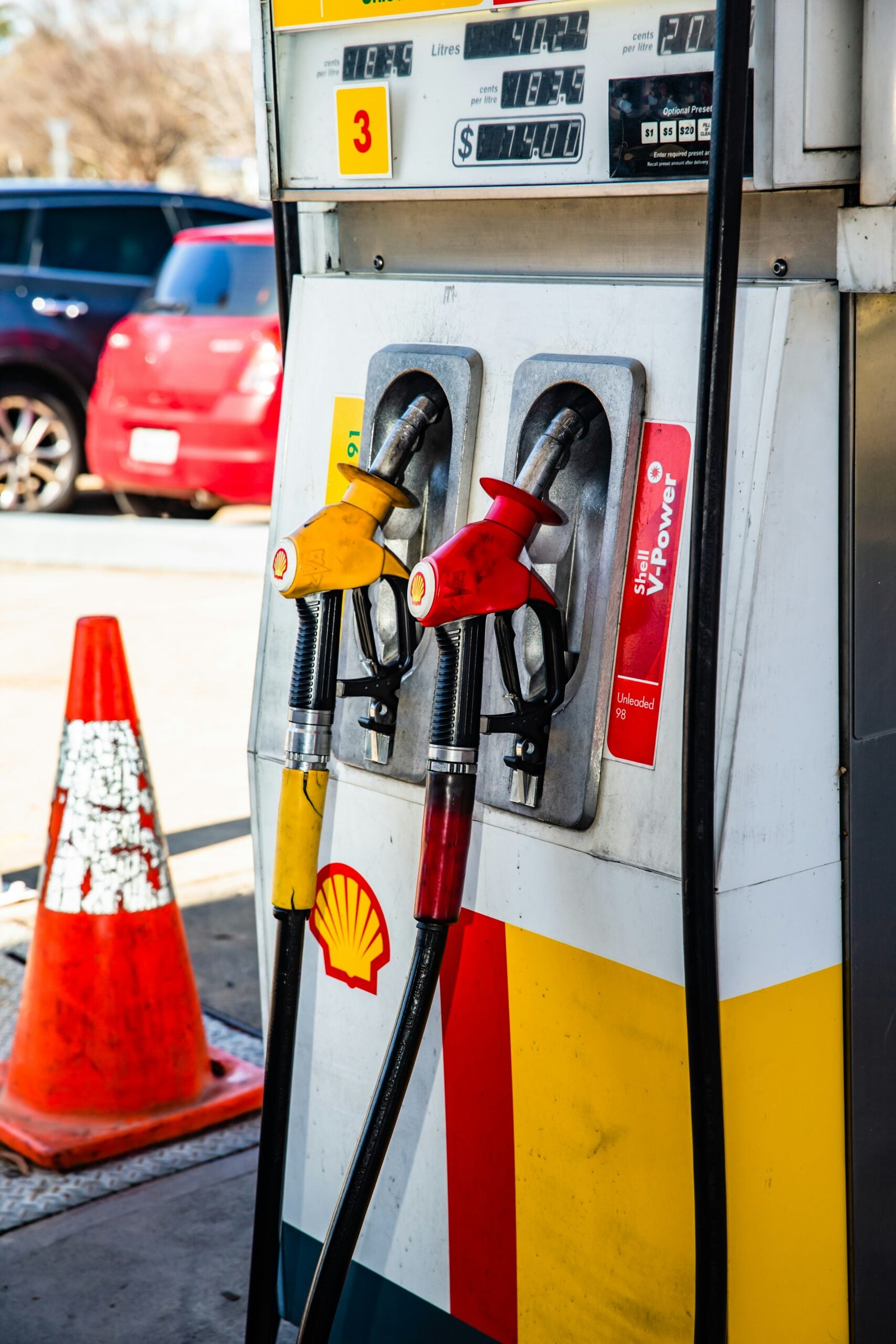A Shell gas pump with two fuel nozzles, one yellow and one red, is pictured next to an orange traffic cone at a gas station. A red SUV and another car are visible in the background.