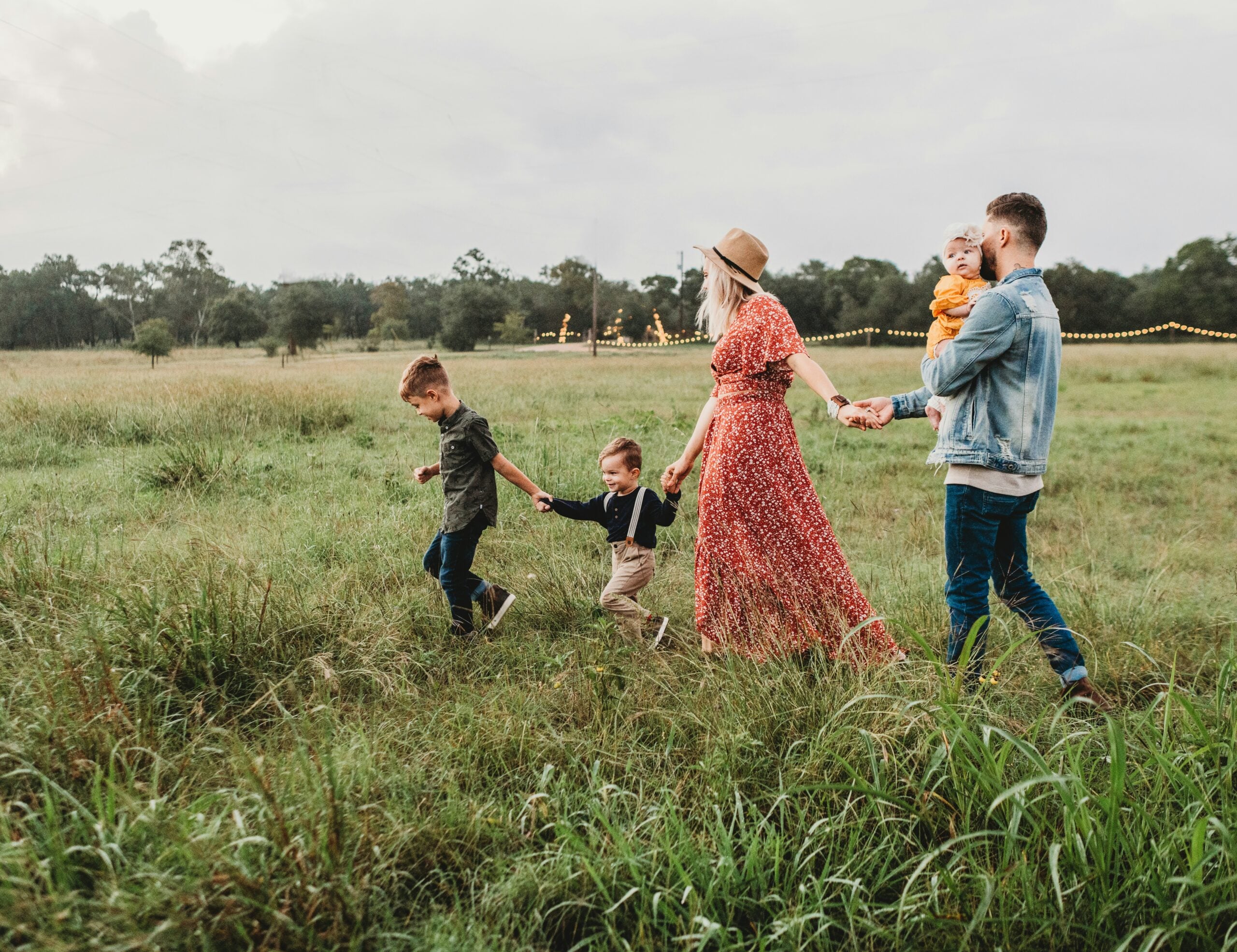 A family of five walks through a grassy field. The mother in a red dress and hat holds hands with two young boys, while the father, in a denim jacket, carries a toddler and holds the mother's hand. Trees and lights are visible in the background.