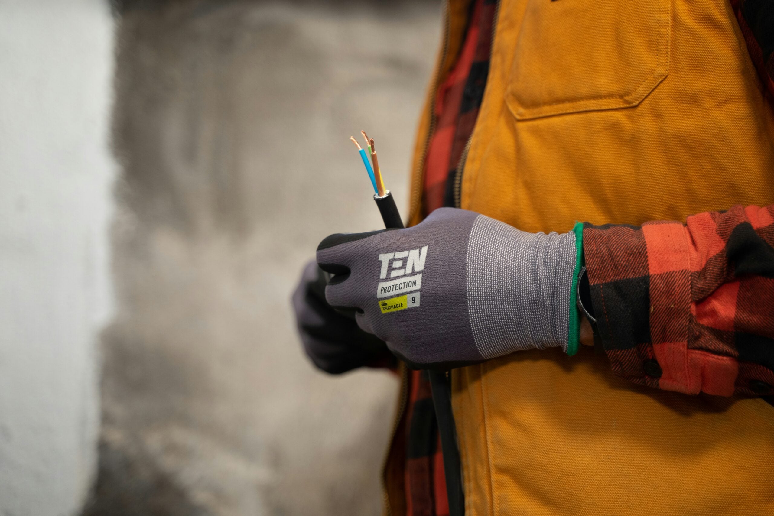 A person wearing gray protective gloves and an orange vest holds a cut electrical wire with exposed copper ends. The background is blurred, drawing focus to the hands and wire.