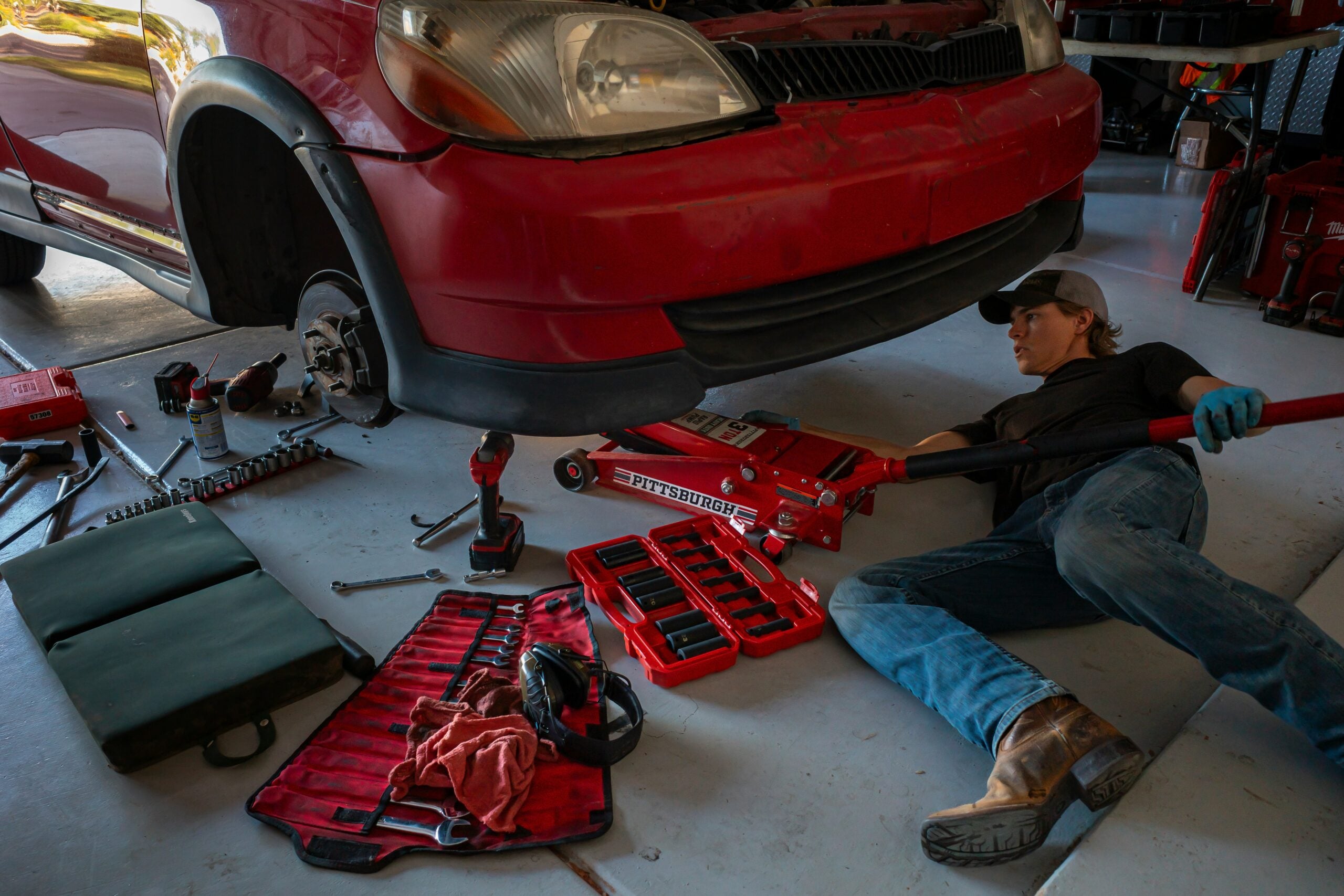 A person lies on the floor using a hydraulic jack to lift a red car with its front wheel removed. Various tools and toolkits are spread out on the garage floor around them.