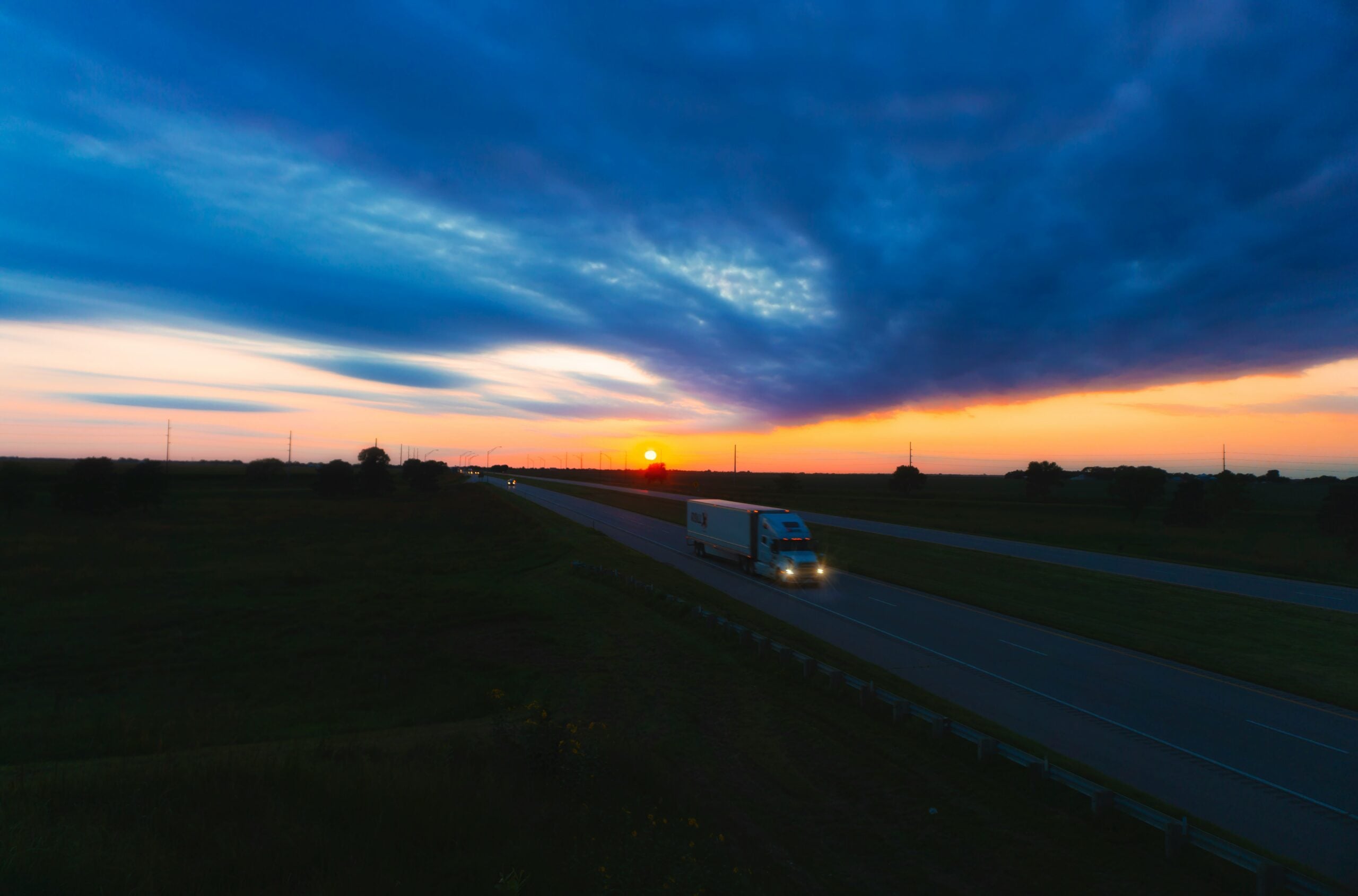A semi-truck drives down a highway at dusk under a dramatic, cloudy sky with the sun setting on the horizon, casting orange, yellow, and blue hues across the landscape.
