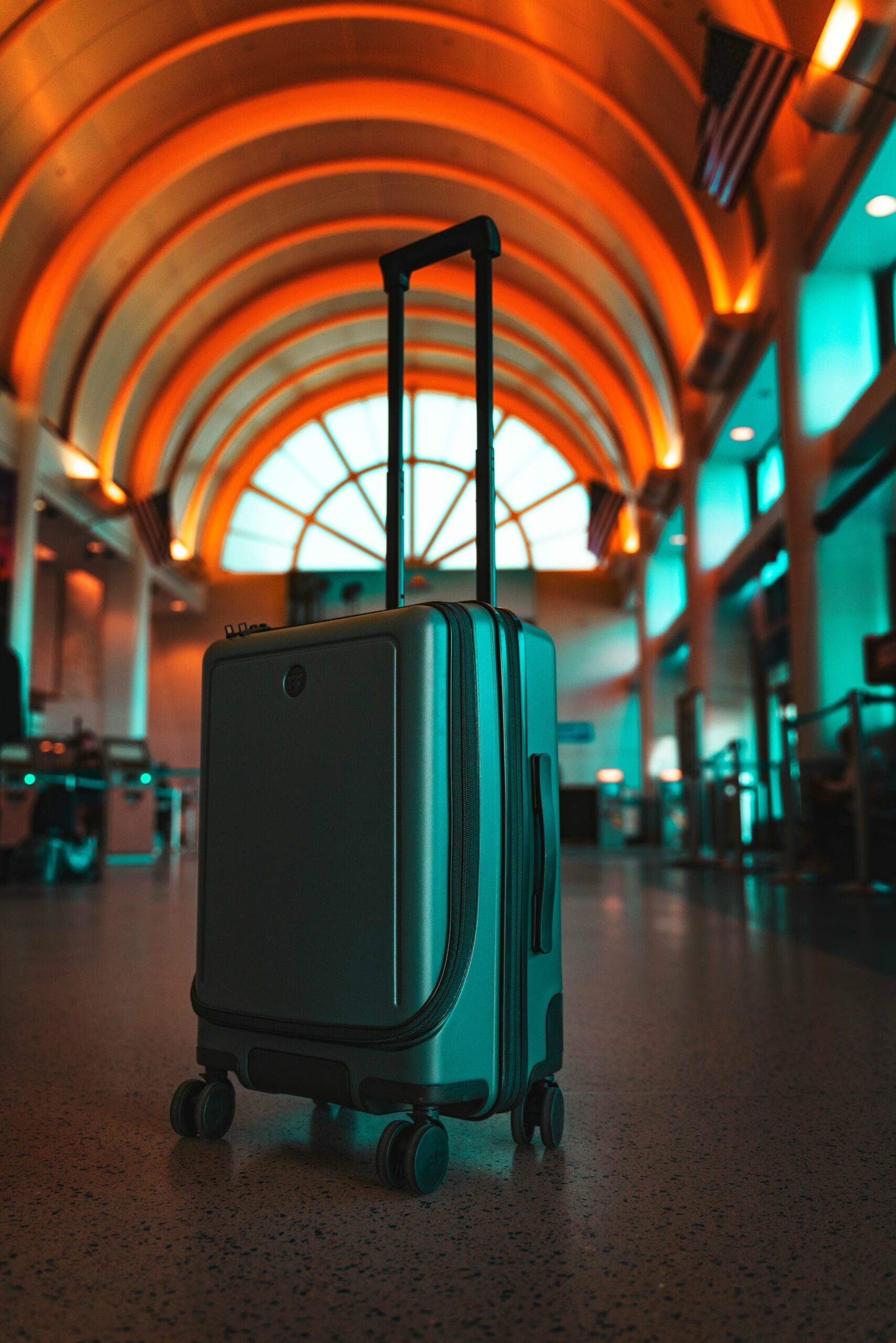A hard-shell suitcase with an extended handle stands alone in a modern airport terminal with a curved, illuminated ceiling and American flags hanging above.