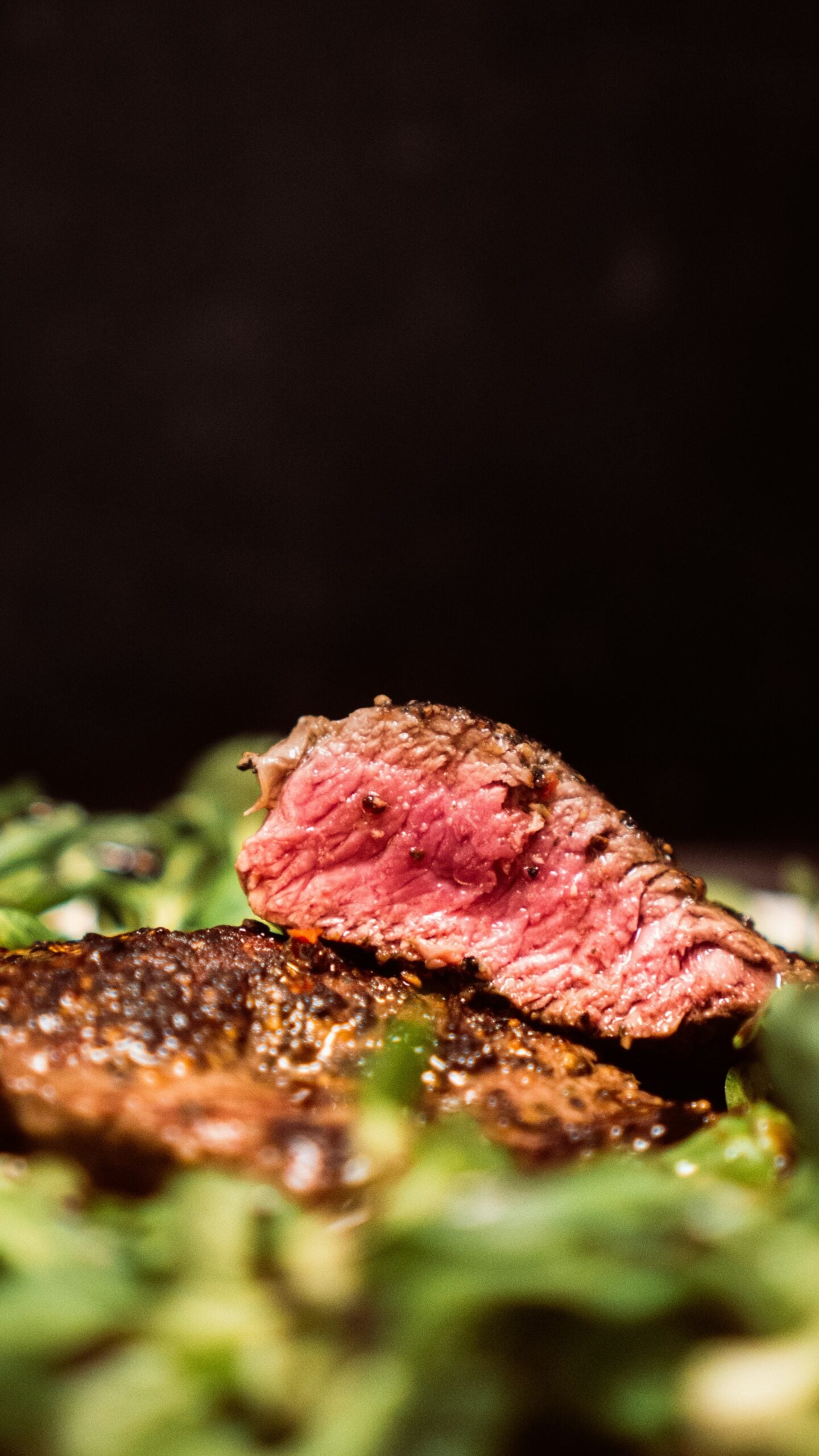 A close-up of a sliced medium-rare steak, showing a juicy pink center with a seasoned, seared crust, resting on a bed of green leafy vegetables against a dark background.