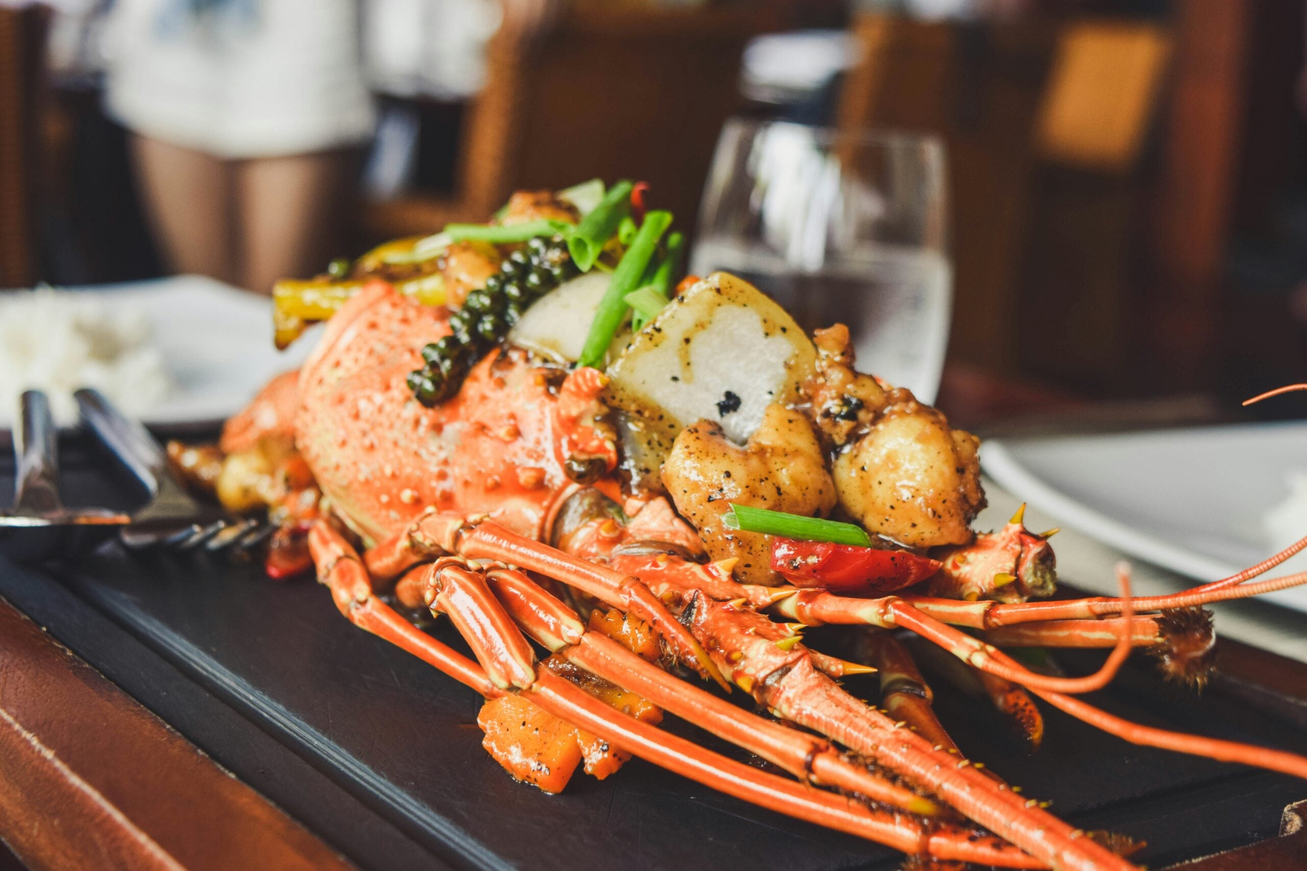 A close-up of a cooked lobster served on a black platter, garnished with green onions and vegetables, with a glass of water and cutlery in the background on a restaurant table.