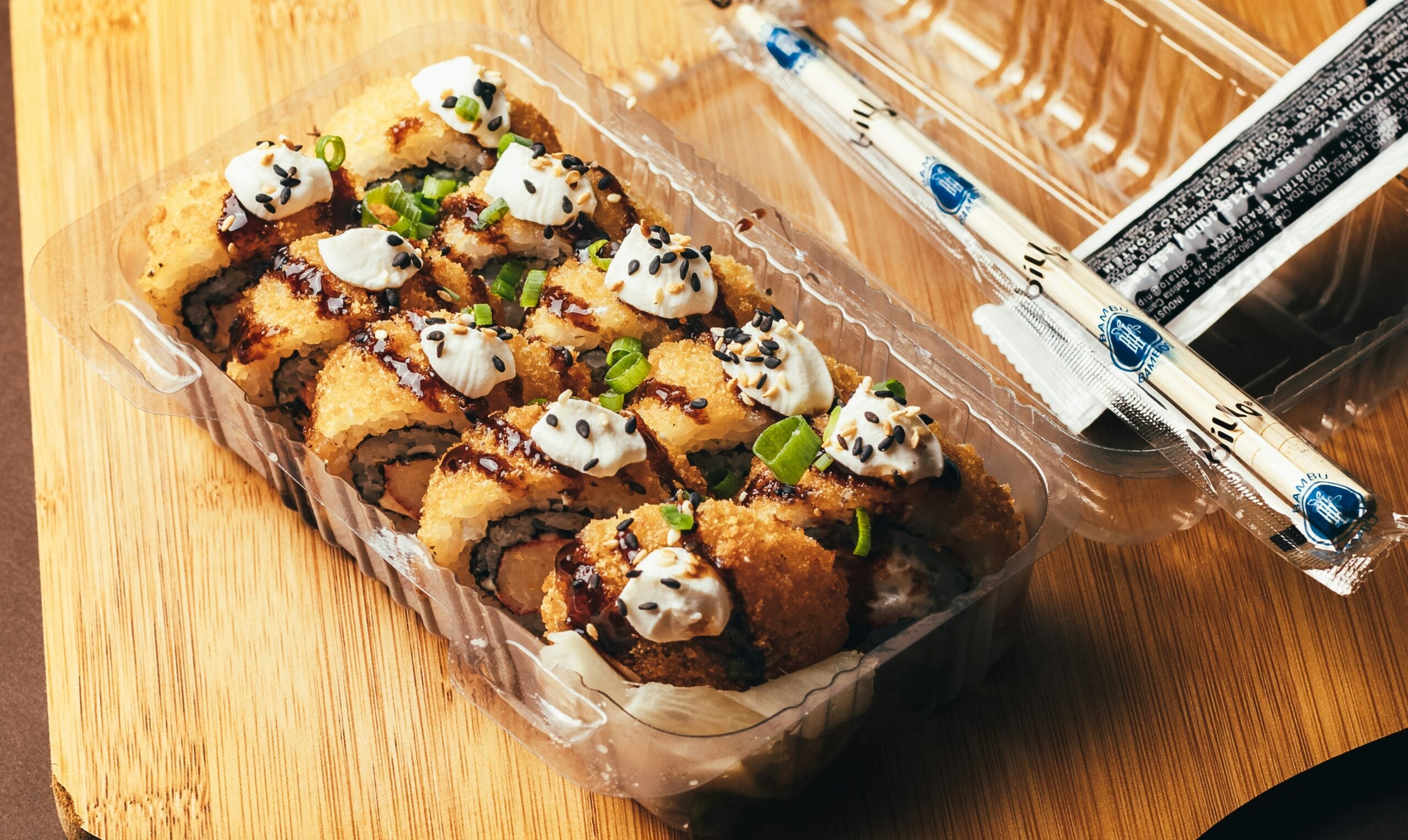 A clear plastic container with sushi rolls topped with sauce, cream, sesame seeds, and green onions, placed on a wooden surface. Chopsticks and a soy sauce packet are beside the container.