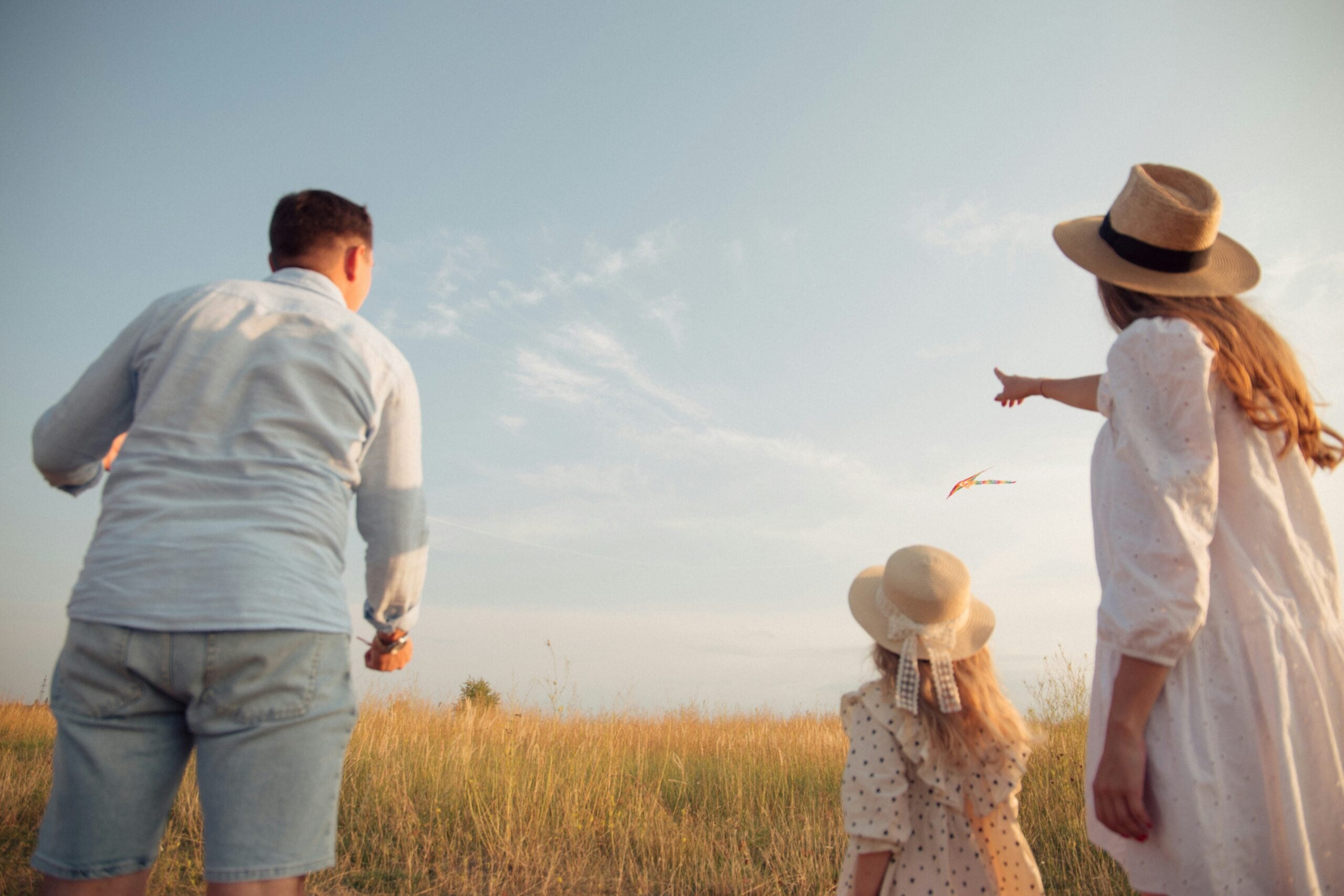 A family of three, seen from behind, stands in a grassy field. The mother points toward a colorful kite flying in the sky, while the father and young daughter watch. All wear light-colored clothes and summer hats.