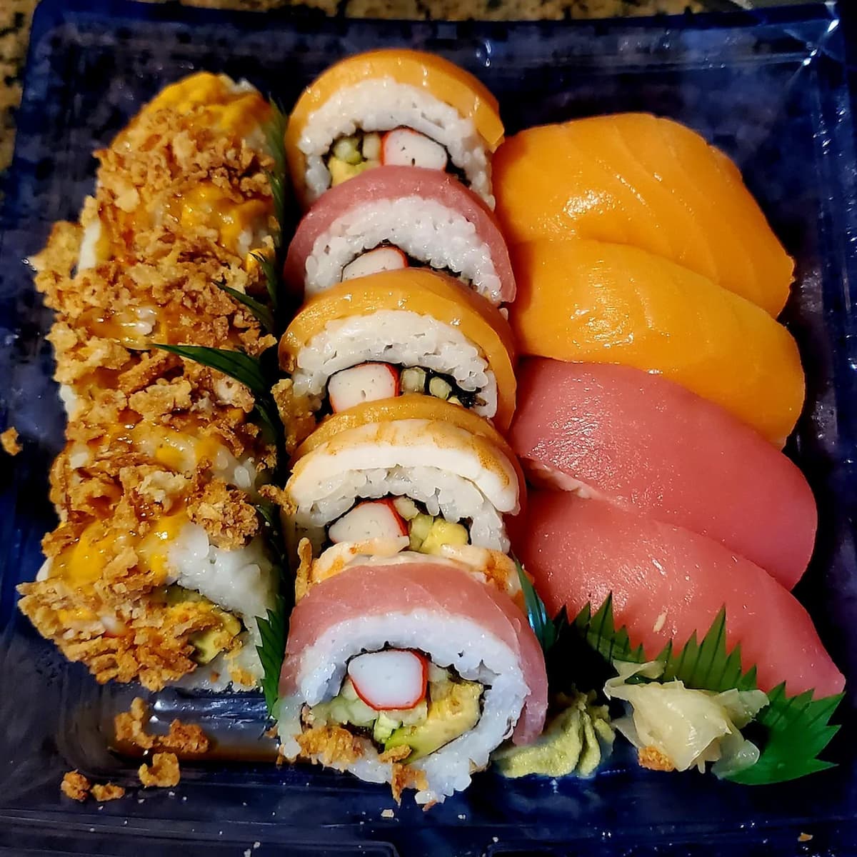 A blue plastic container holds a variety of sushi rolls and nigiri. The rolls feature white rice, imitation crab, and either orange or pink fish. One roll is covered in crispy fried onions and a yellow sauce.