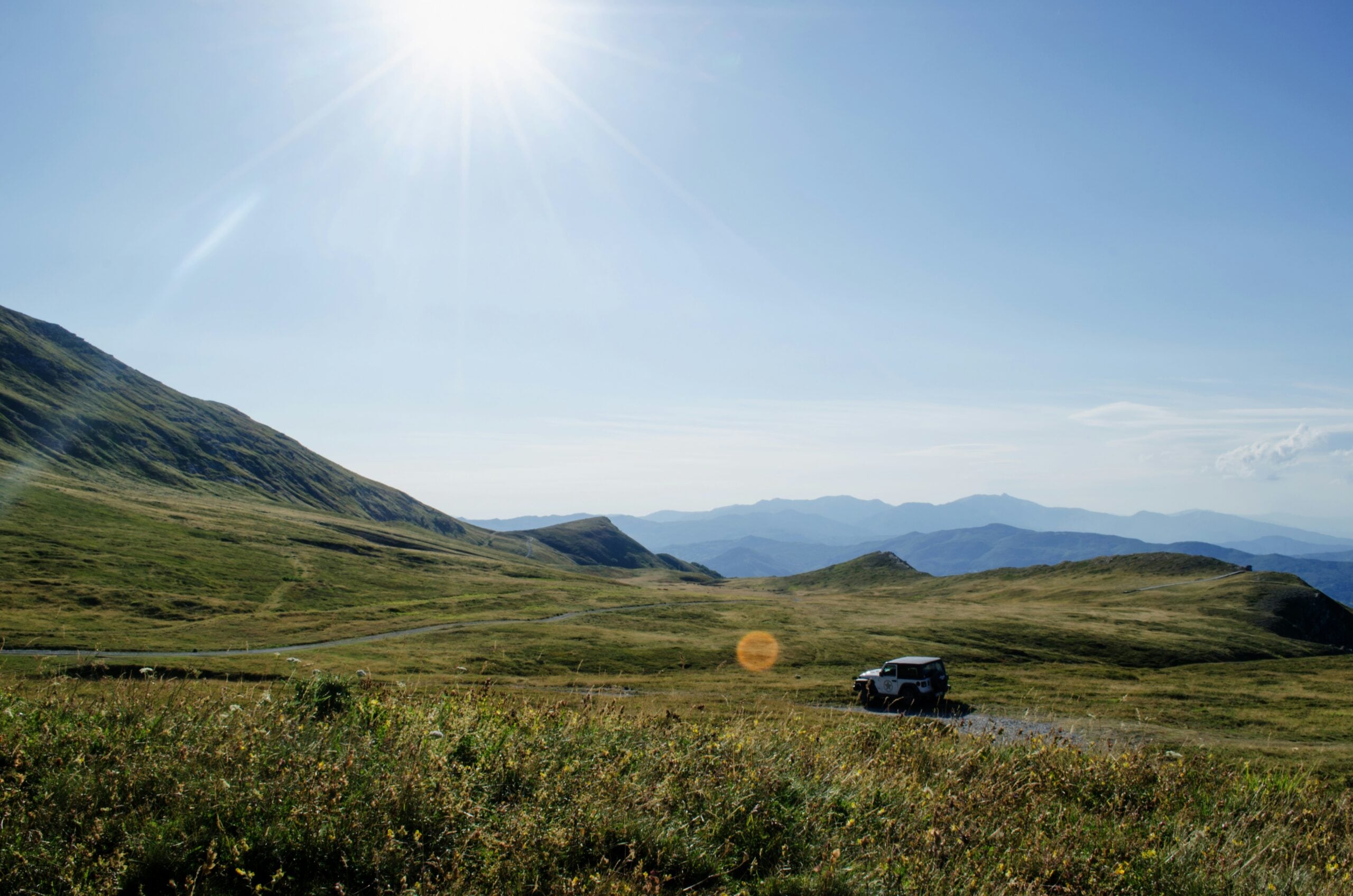 A car drives along a winding road through green, rolling hills under a bright sun and clear blue sky, with distant mountain ranges in the background.