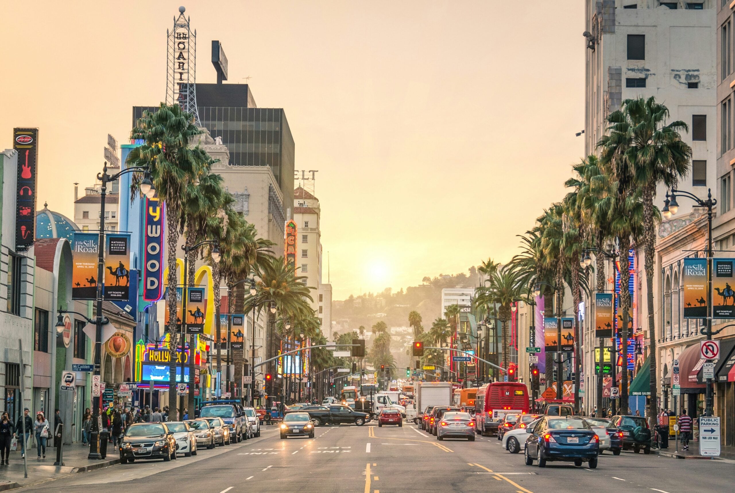 Busy street scene on Hollywood Boulevard at sunset, with palm trees, cars, people on sidewalks, and colorful billboards and neon signs for theaters and shops lining both sides of the street. Most expensive and cheapest states to travel.