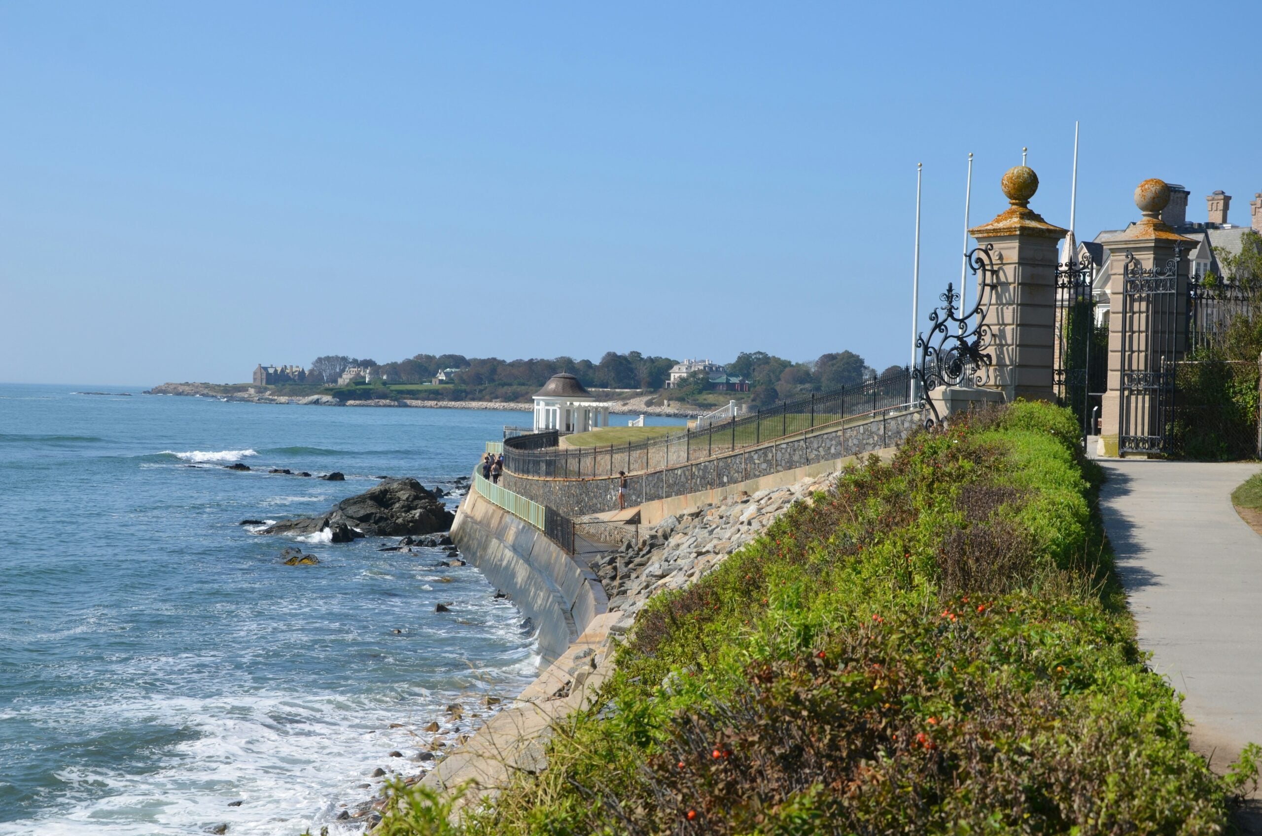 A seaside walkway runs beside a tall iron gate with stone pillars, lush green bushes, and ocean waves crashing against a rocky shoreline under a clear blue sky.