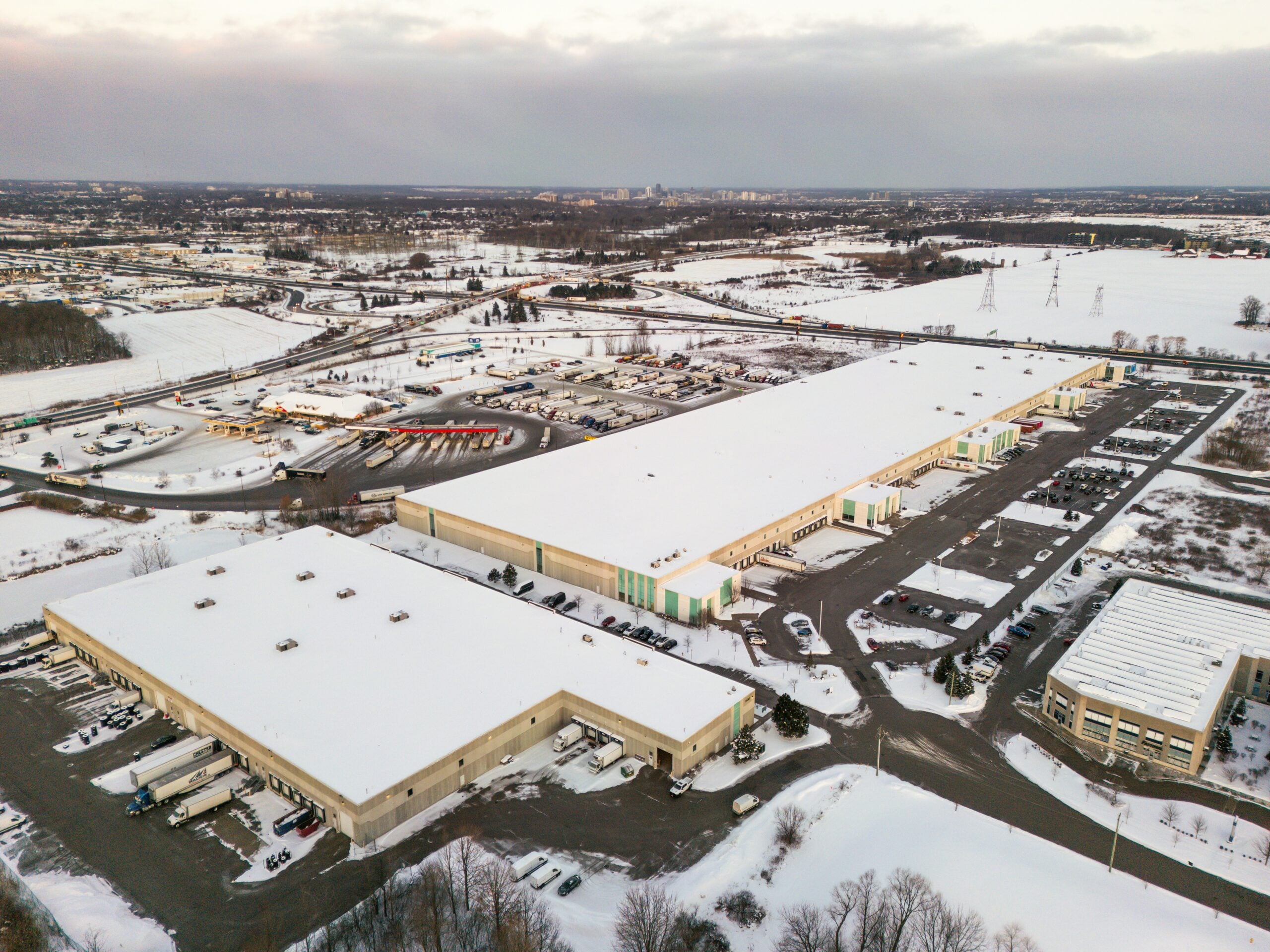 Aerial view of large industrial warehouses and parking lots covered in snow, surrounded by roads, vehicles, and open fields on a cloudy winter day.