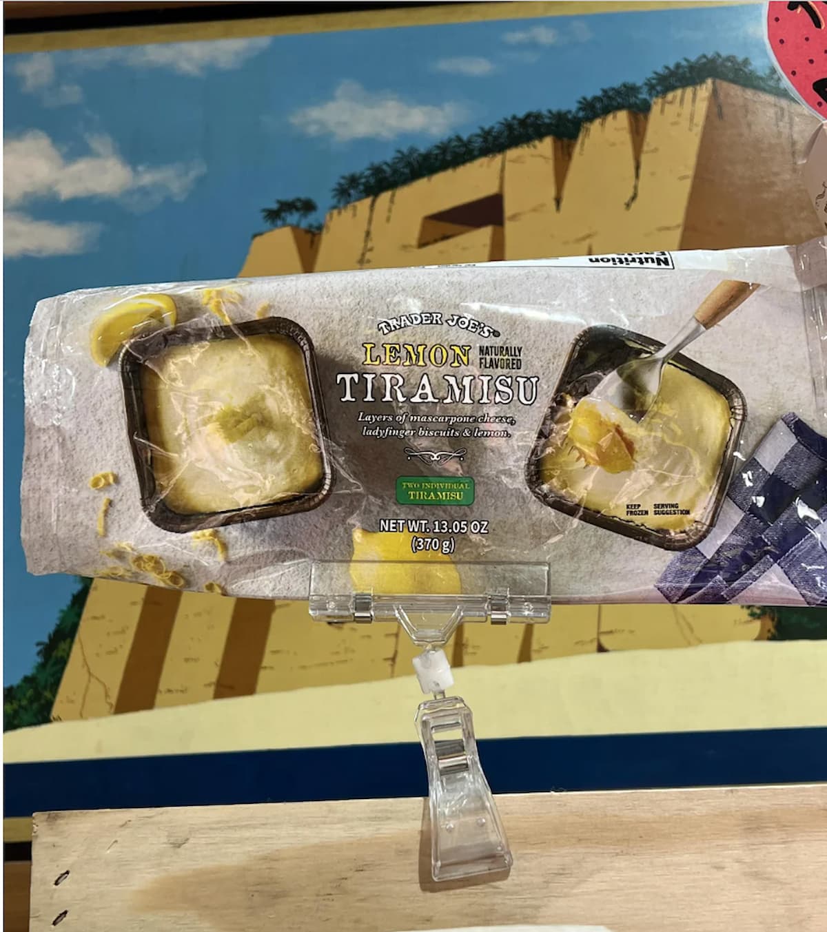 A package of Trader Joe's Lemon Tiramisu is clipped to a display shelf, with a colorful painting of a cliff and trees in the background. The package shows images of the dessert and text highlighting the lemon flavor.