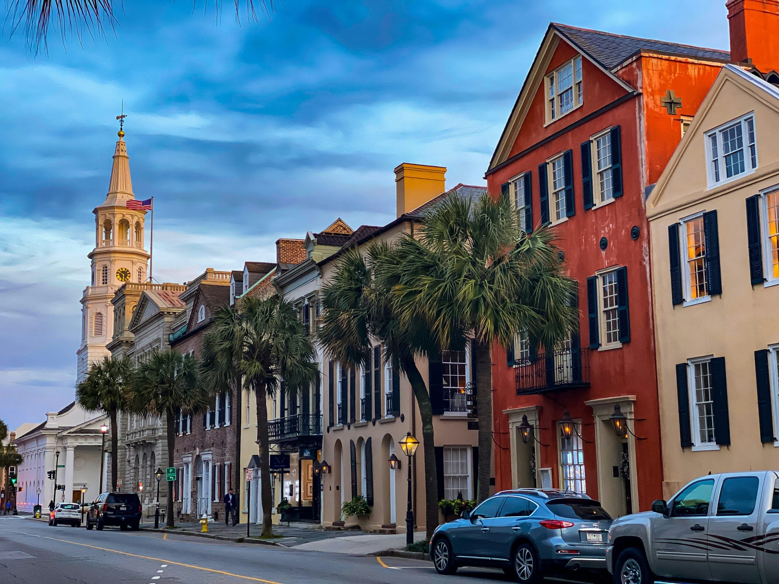 A row of colorful historic buildings and palm trees line a street at sunset, with parked cars and a church steeple visible in the background under a partly cloudy sky.
