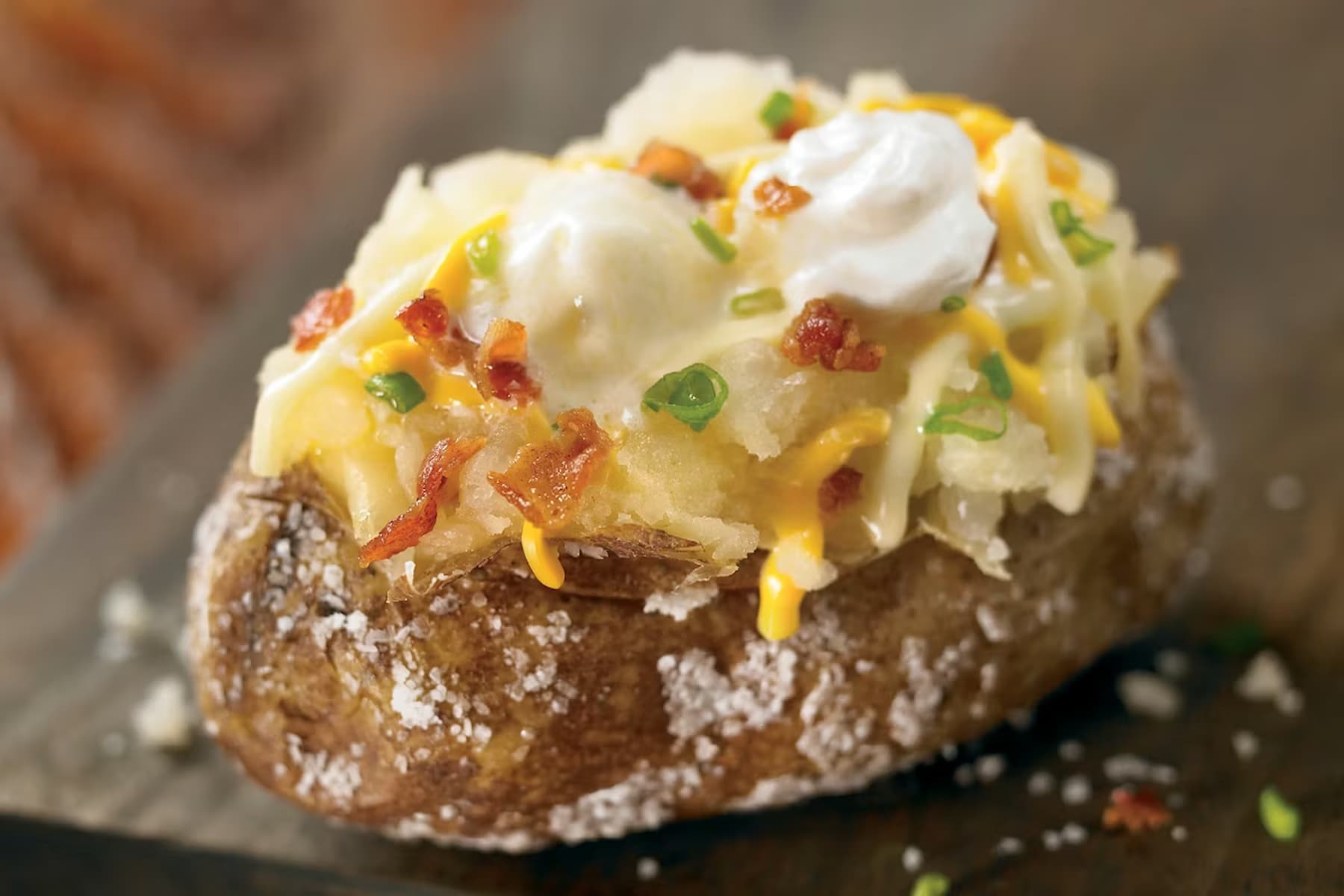 A baked potato topped with melted cheese, sour cream, crumbled bacon, and chopped green onions, served on a wooden surface.