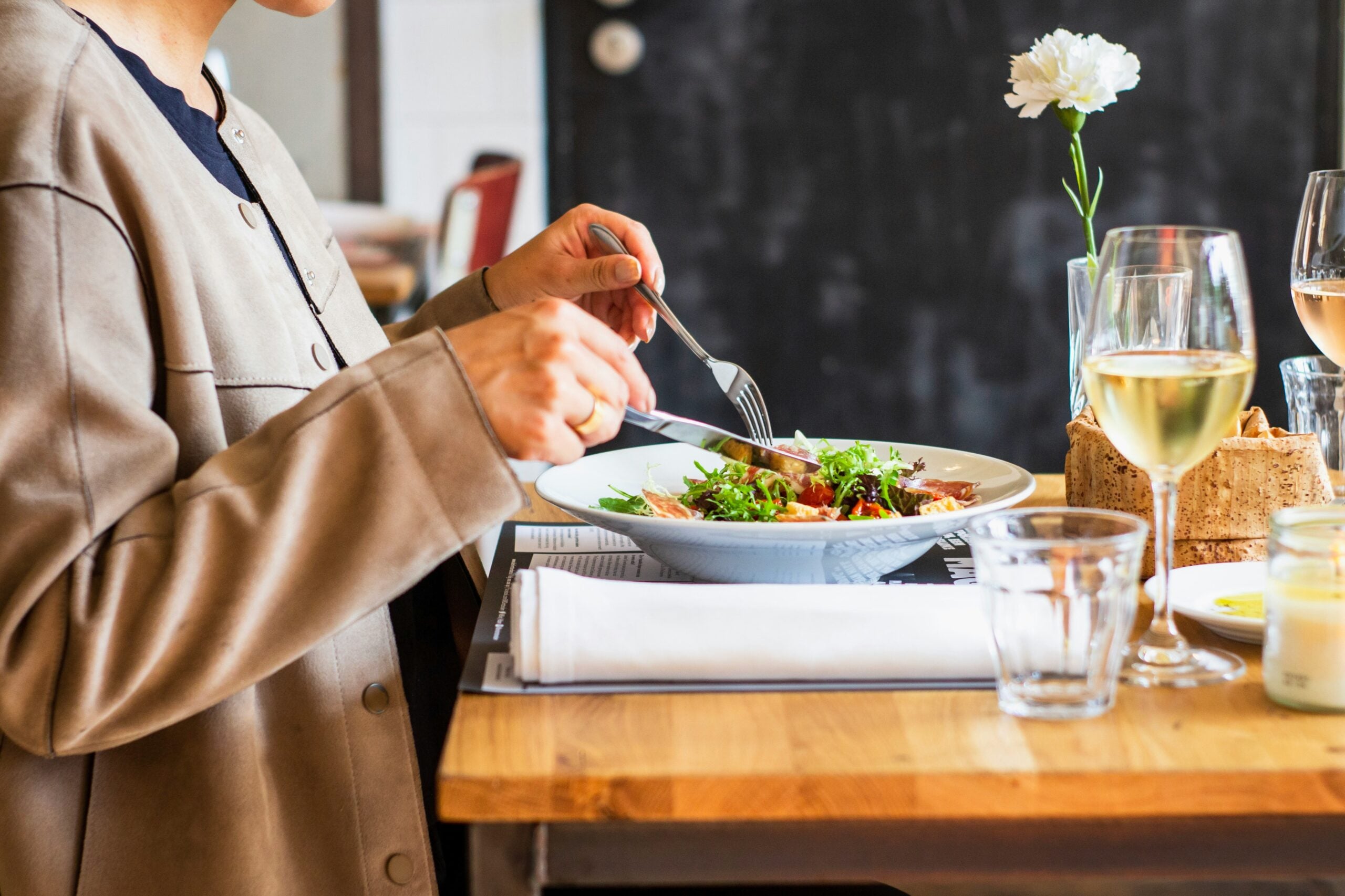 A person sits at a wooden table, eating a salad with a fork and knife. There are glasses of white wine and water, a flower in a vase, and bread on the table. The setting appears to be a restaurant.