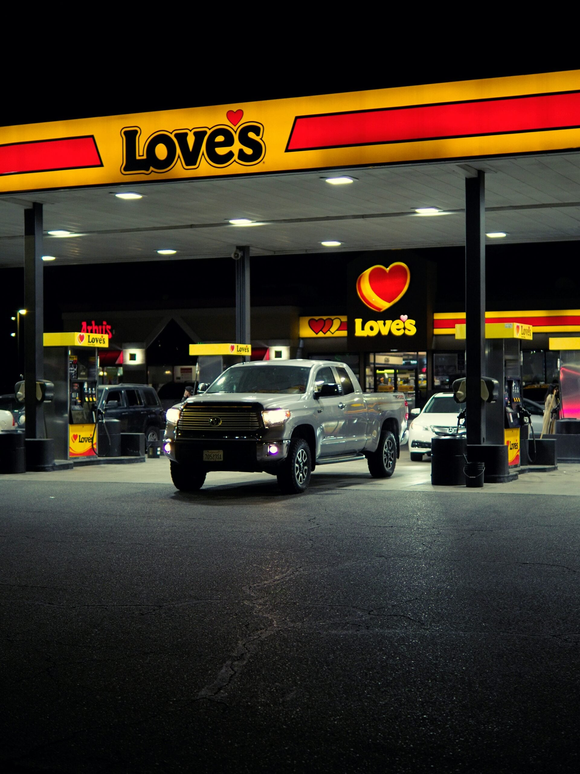 A silver pickup truck is parked under the bright yellow and red canopy of a Love’s gas station at night, with the Love’s logo prominently displayed and other vehicles in the background.