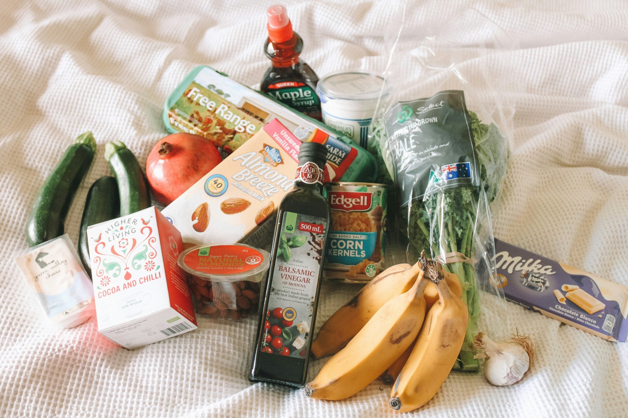 A variety of groceries on a white surface, including bananas, zucchini, pomegranate, spinach, kale, maple syrup, almond breeze milk, corn, tea, balsamic vinegar, garlic, and a Milka chocolate bar.
