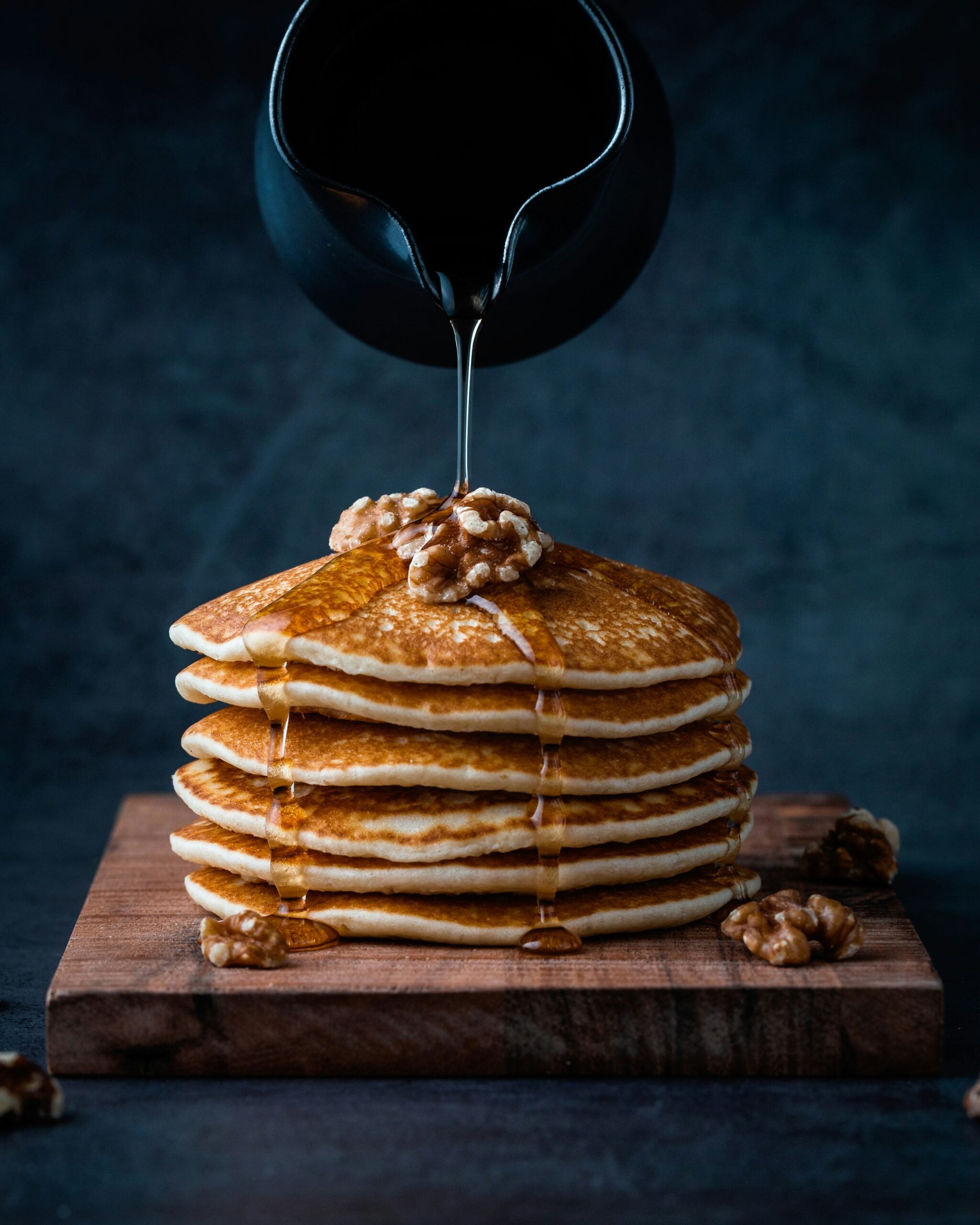 A stack of pancakes topped with walnuts is drizzled with syrup from a black pitcher, set on a wooden board against a dark background.
