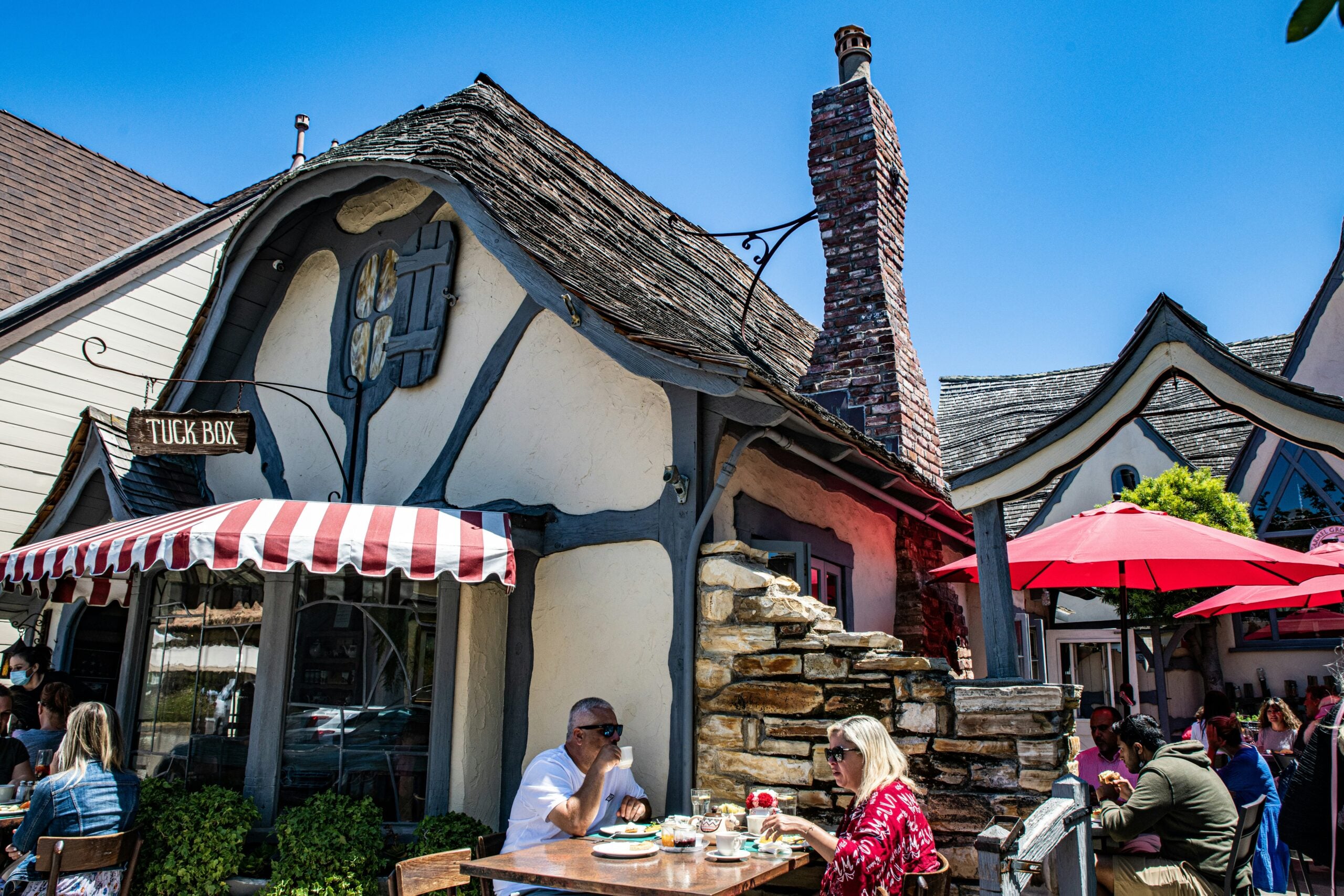 People dine outdoors at rustic wooden tables in front of a whimsical, cottage-style building with a sloped roof, stone chimney, and a sign that reads “Tuck Box.” Red and white umbrellas provide shade on a sunny day.